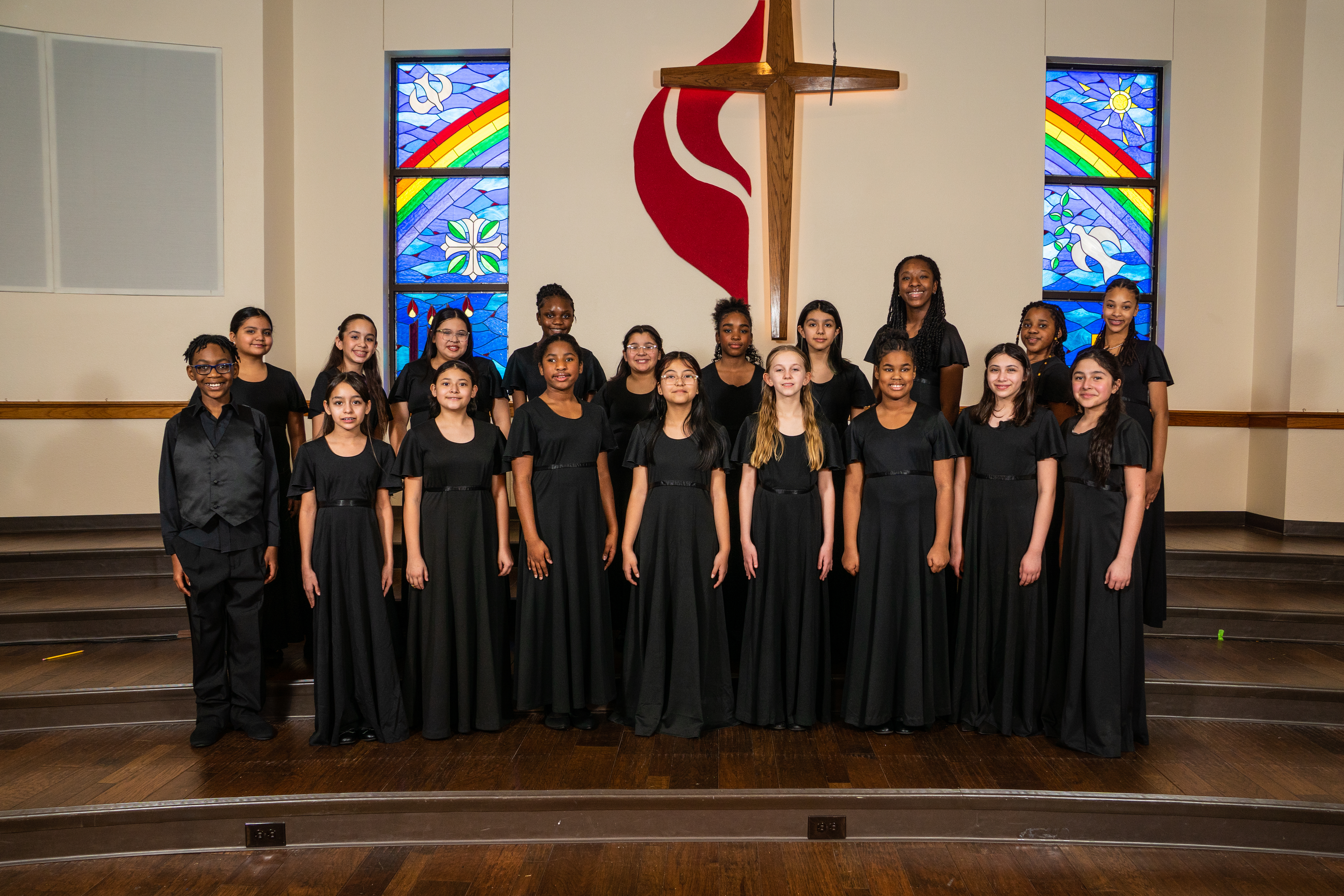 A group of young people, dressed in black, standing in a church-like setting with stained glass windows and a cross in the background.
