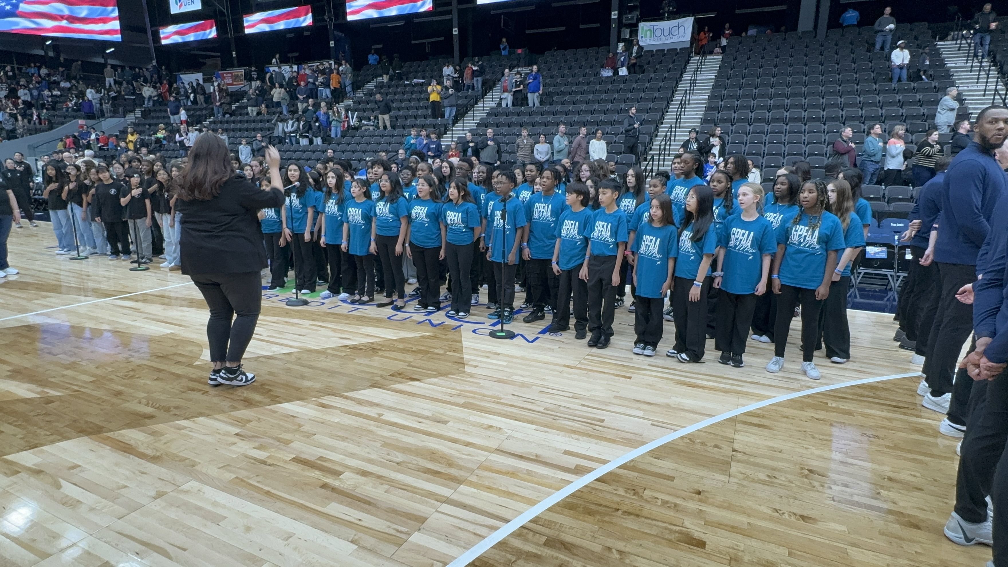 A large group of people in matching blue shirts stand on a basketball court, with an American flag displayed in the background.