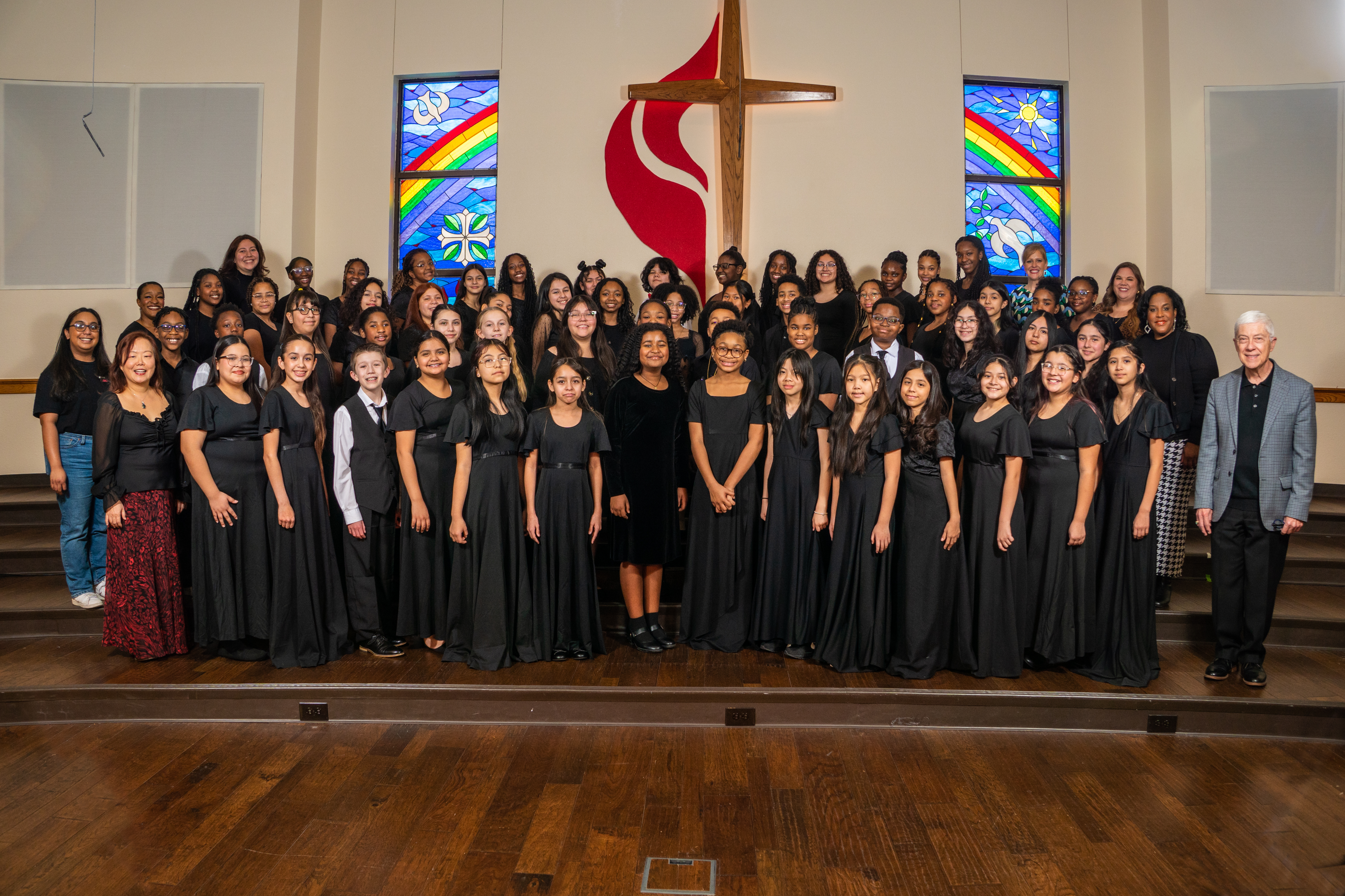 A large group of people, dressed in black, standing in a church-like setting with stained glass windows and a cross in the background.