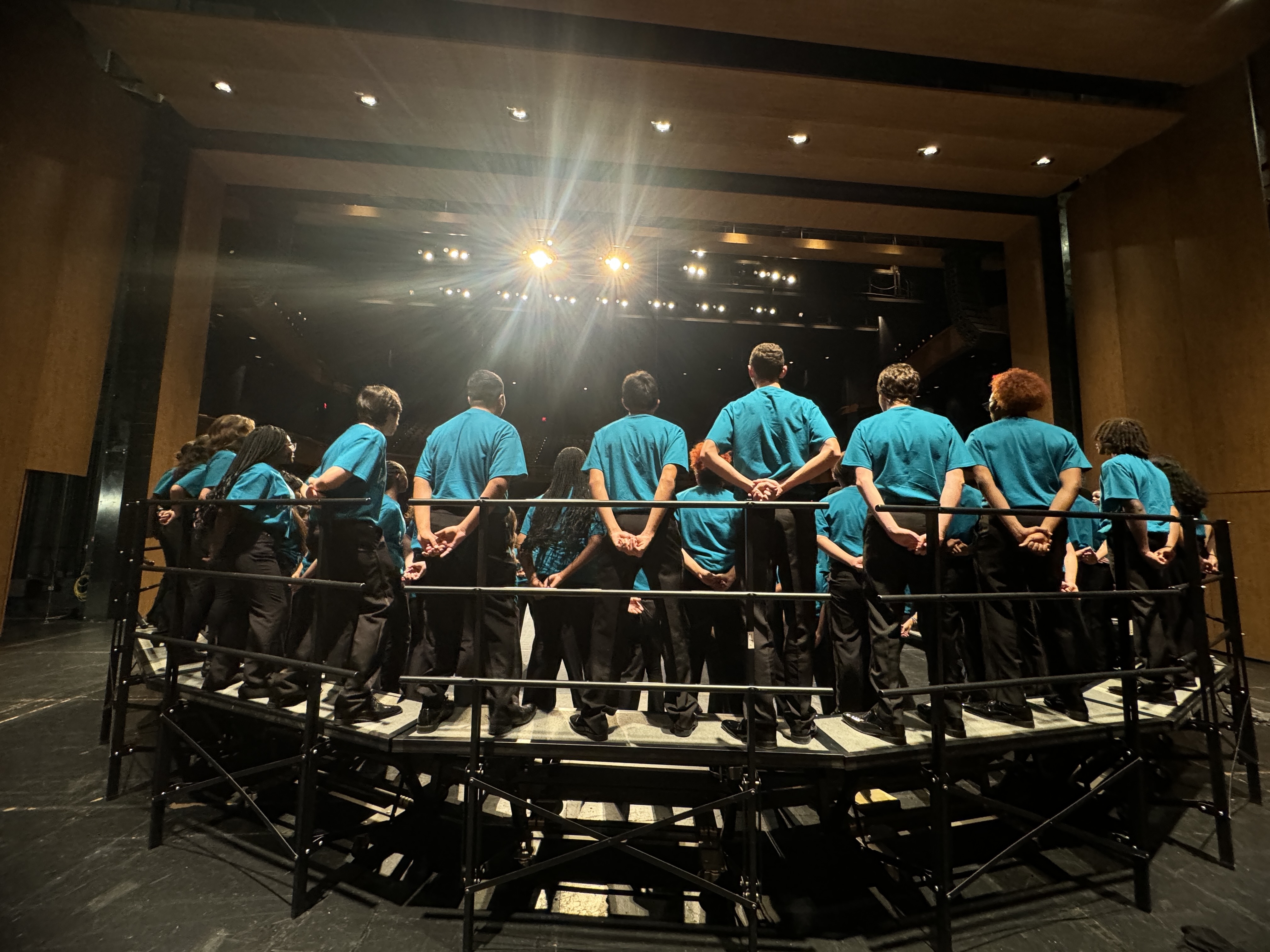 A group of people wearing matching turquoise shirts are standing in a semicircle on a stage with bright lighting and a dark background.