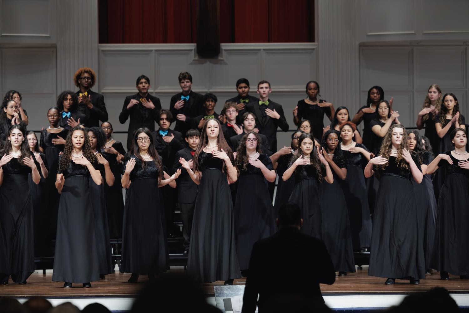 A large group of women dressed in black gowns standing on a stage, with a conductor facing them in the foreground.
