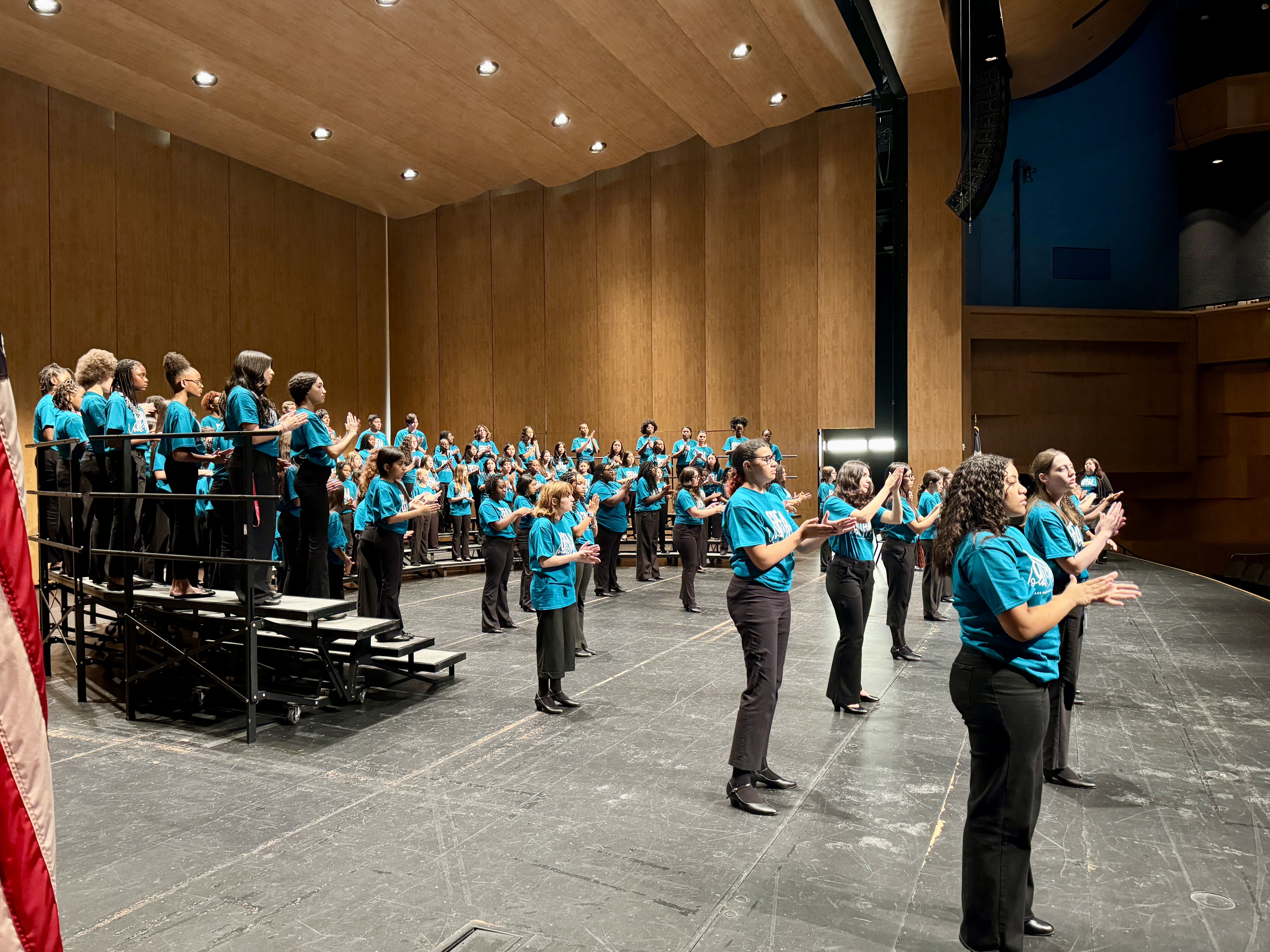 A large group of people in matching turquoise shirts are gathered on a stage in a wooden-paneled auditorium, with a red curtain visible in the background.