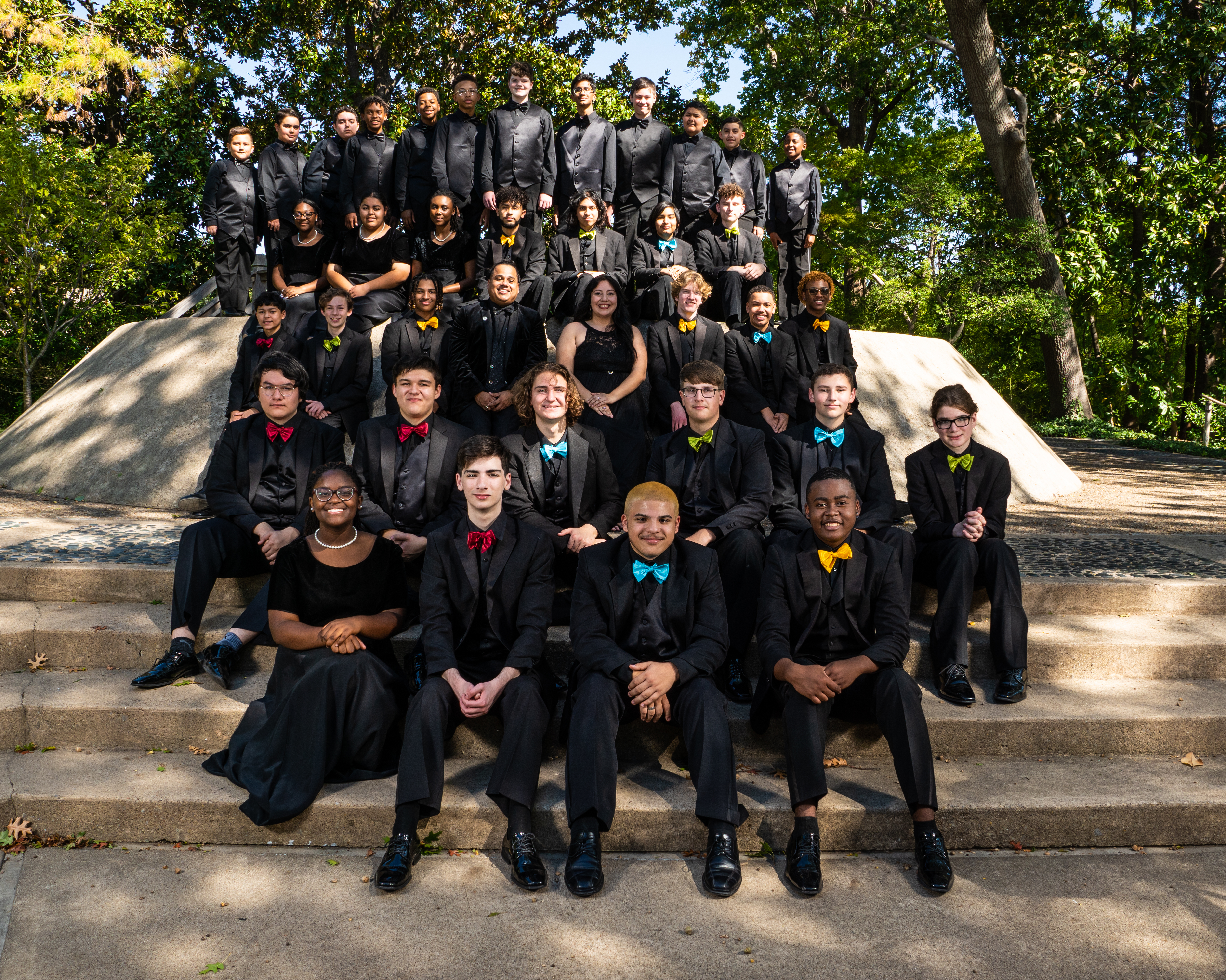 A large group of people, dressed in formal black attire, are gathered together on a set of stone steps surrounded by trees and greenery.