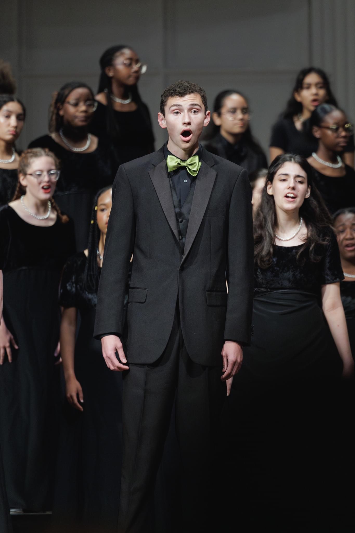 A young man in a formal black suit and bow tie stands in the foreground, surrounded by a group of people dressed in black clothing behind him.