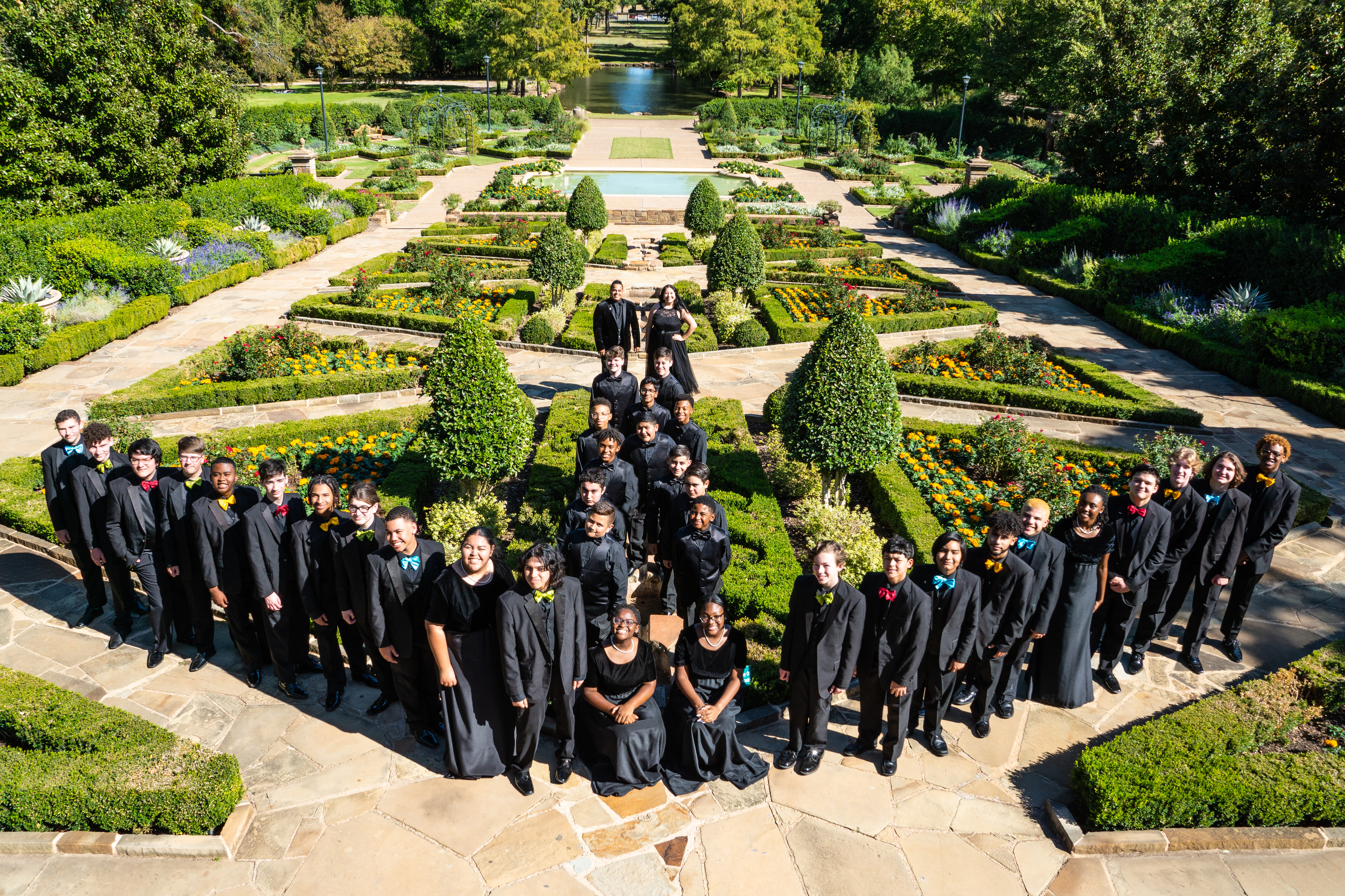 A large group of people dressed in black robes stand in a circular formation in a lush, well-manicured garden with a central water feature and surrounding greenery.
