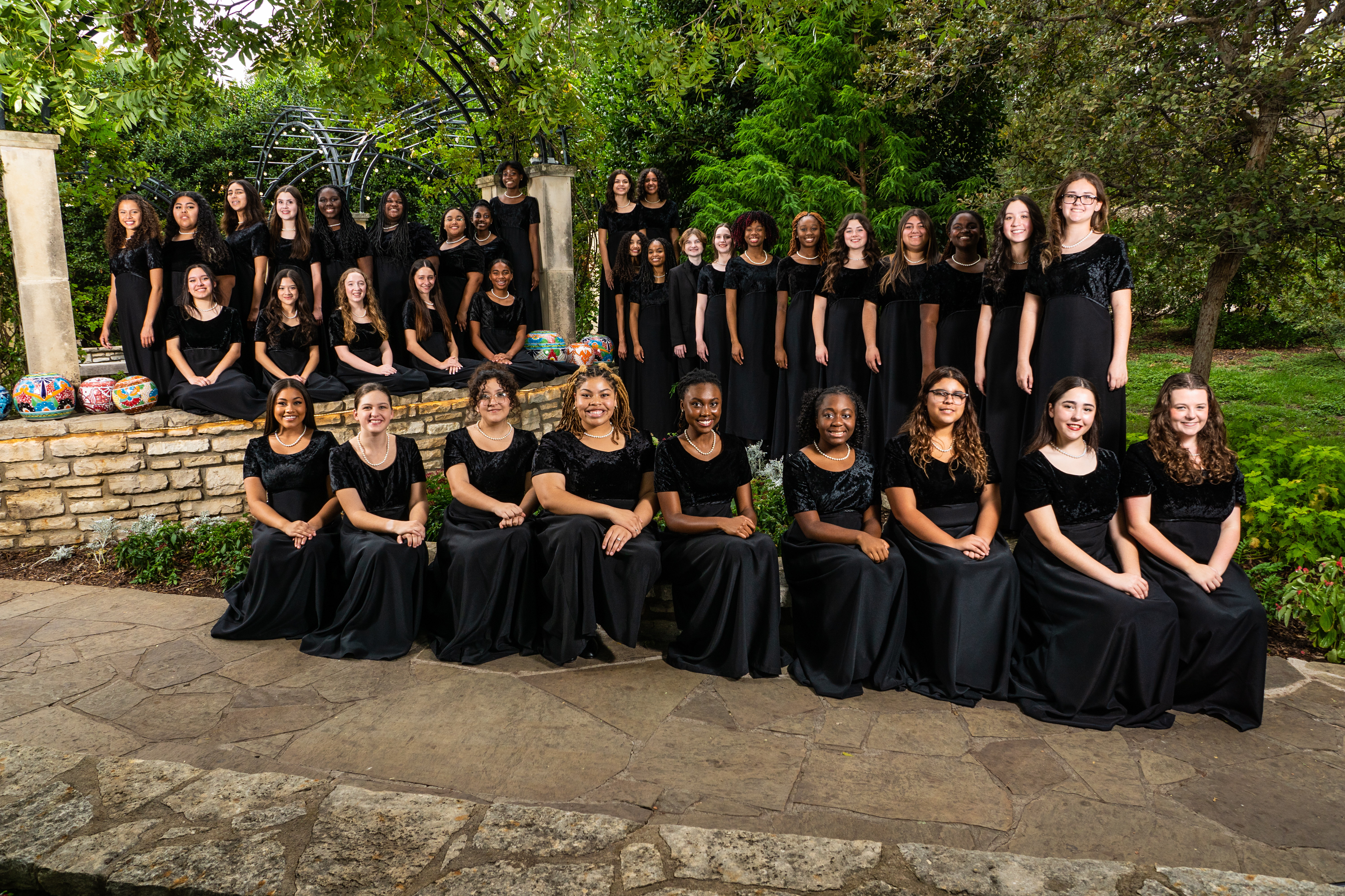A large group of young women dressed in black outfits are gathered together in an outdoor setting with lush greenery and a stone structure in the background.