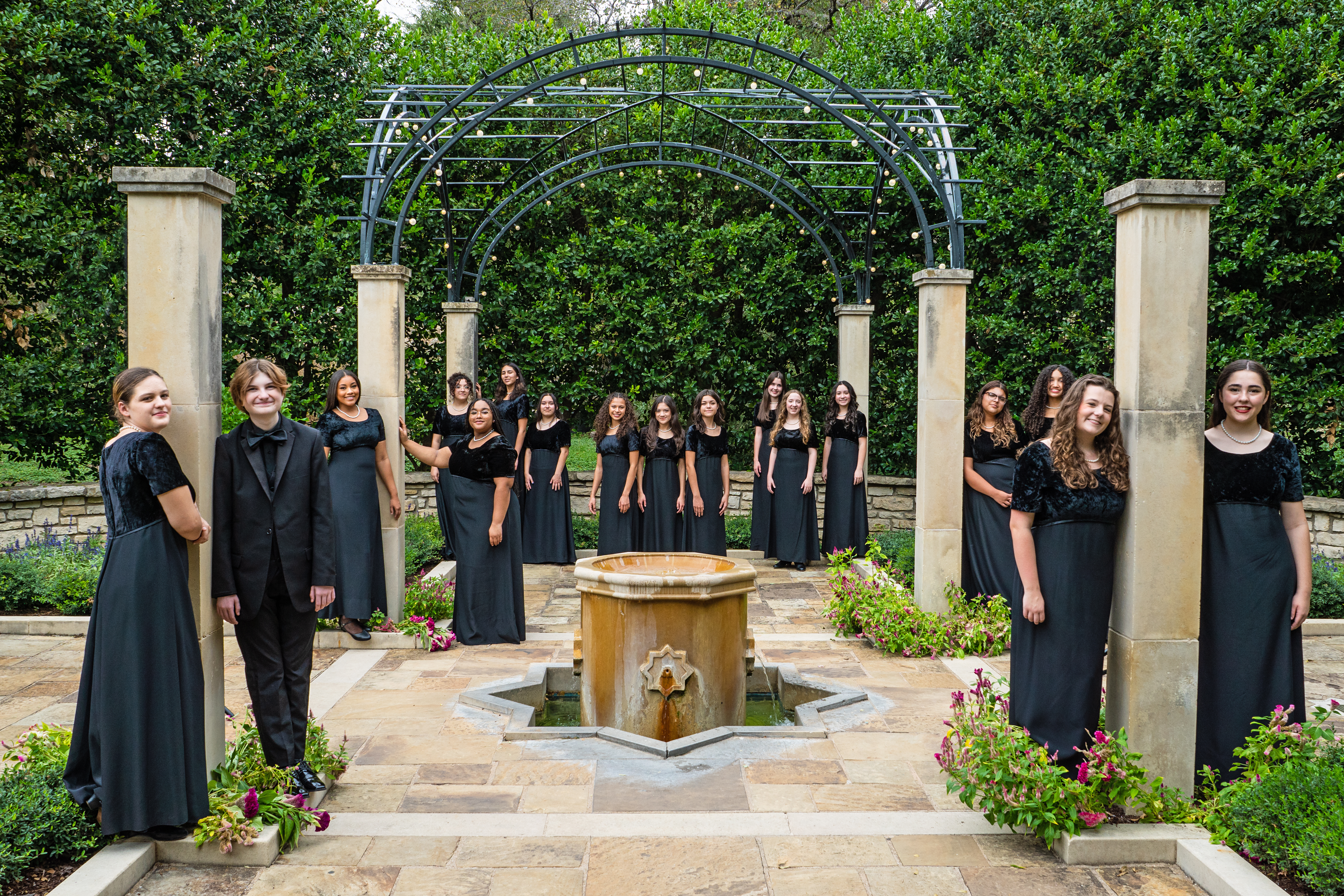 A group of people dressed in black gowns standing in front of an ornate archway surrounded by lush greenery and a fountain in the background.