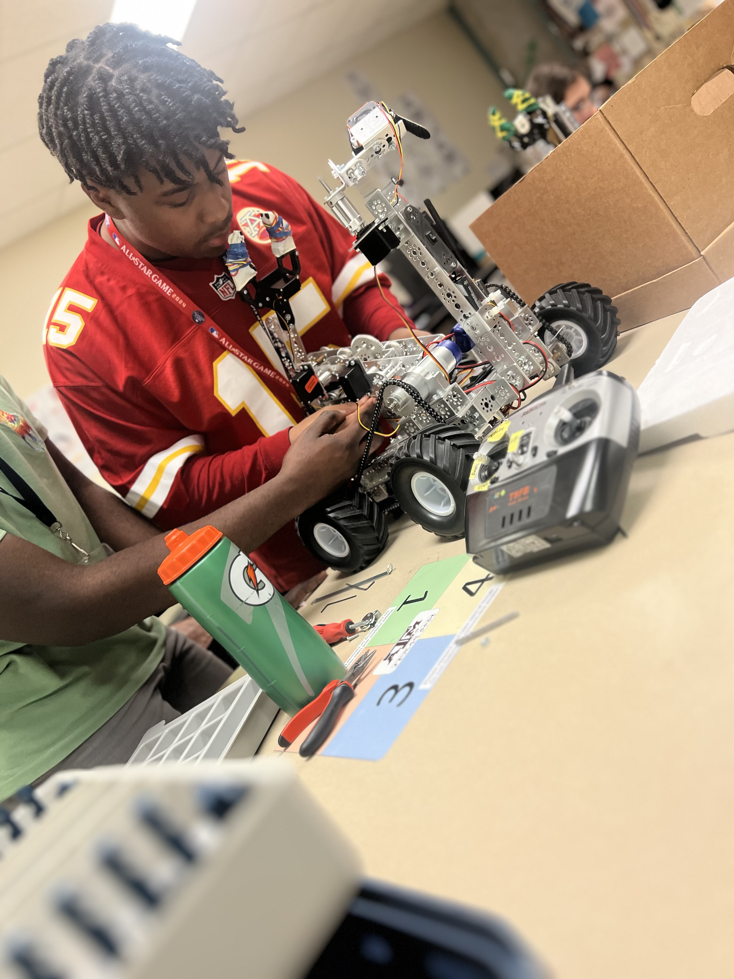 A young person in a red jersey is intently assembling a robotic device on a table, surrounded by various tools and materials in a classroom or workshop setting.
