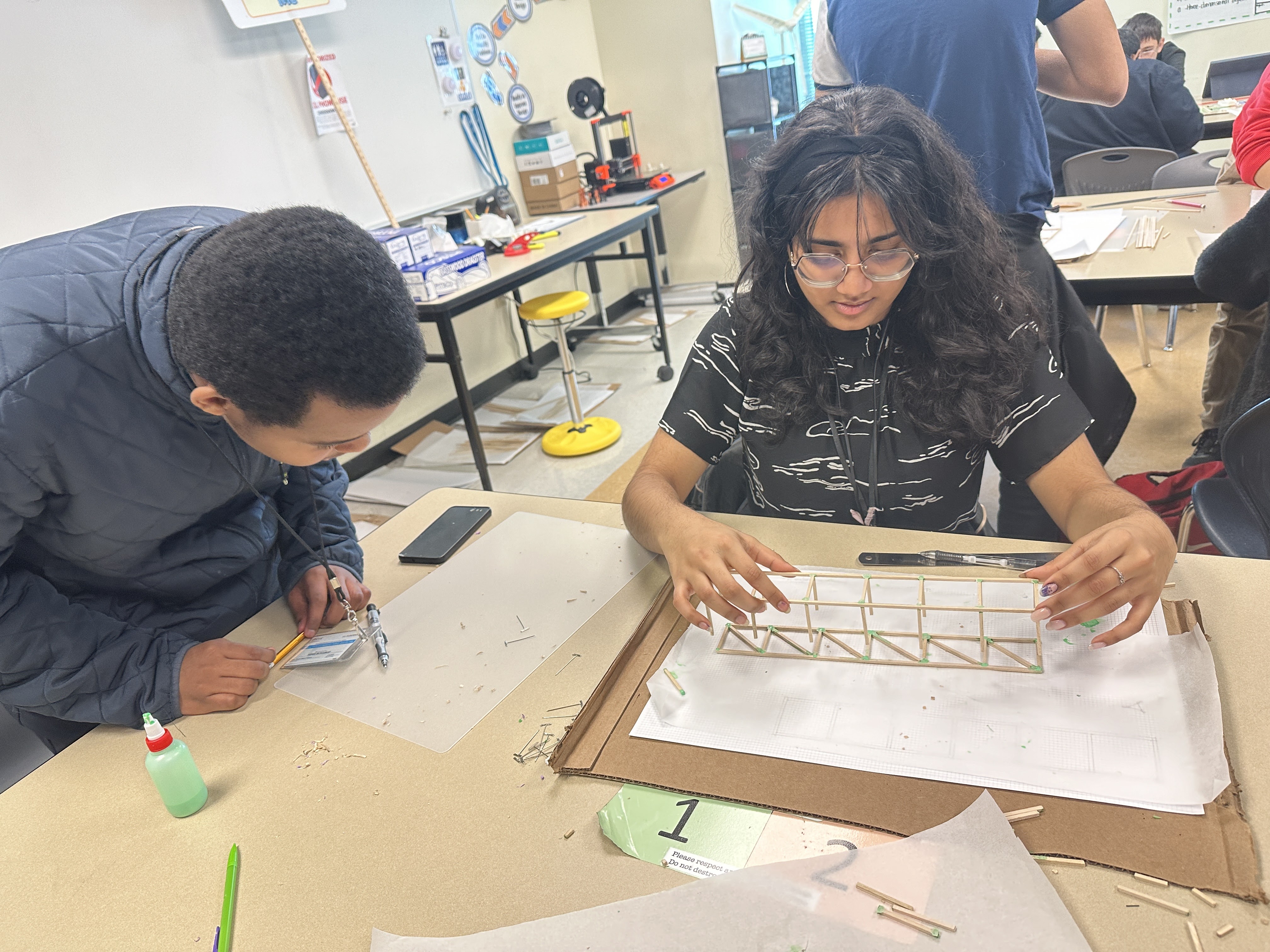 Two individuals, a man and a woman, are working together on a project at a table in what appears to be a classroom or workshop setting, with various supplies and materials visible in the background.