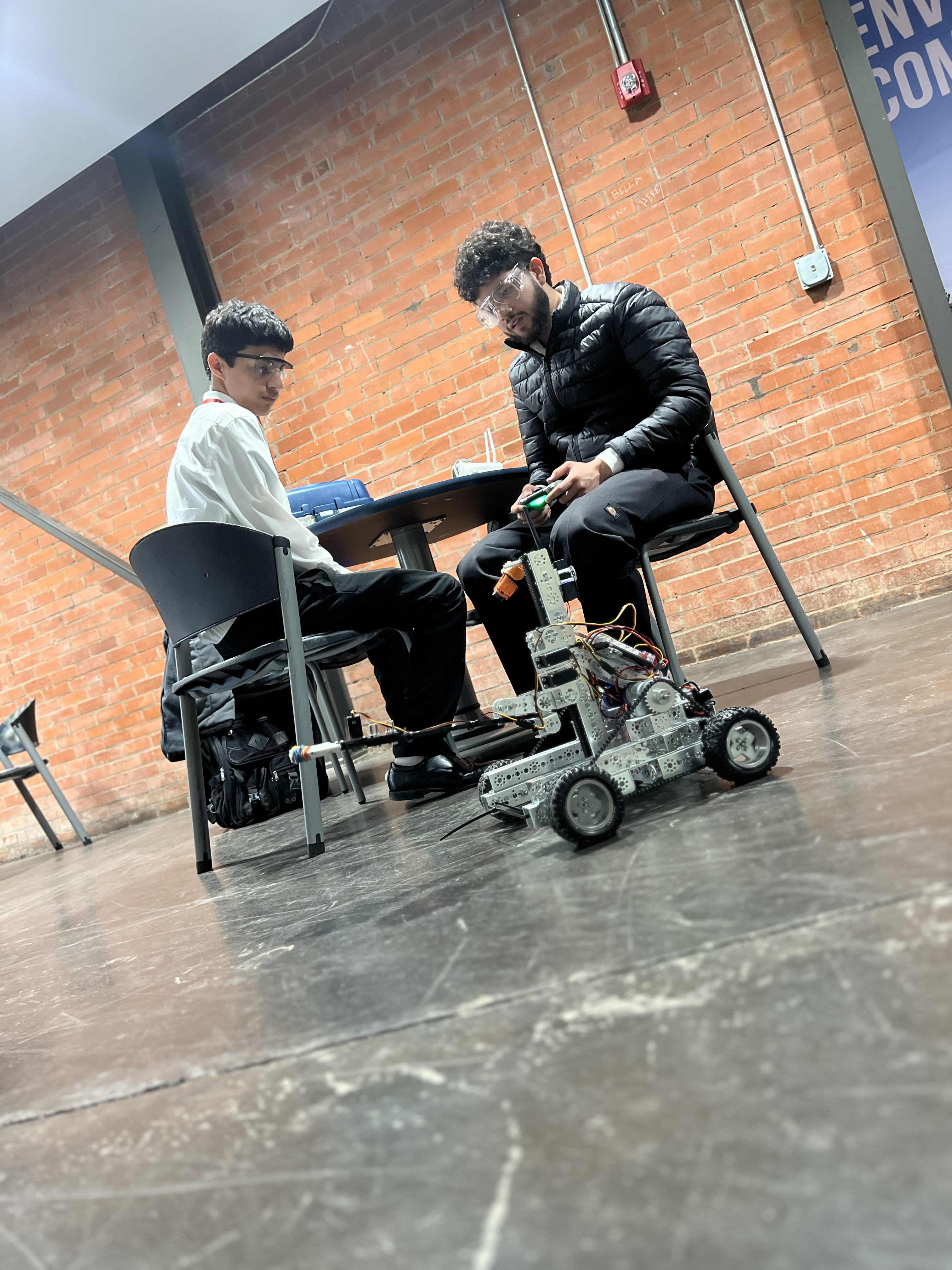 Two individuals, one seated in a wheelchair and the other standing, are engaged in a conversation in a room with a brick wall background.