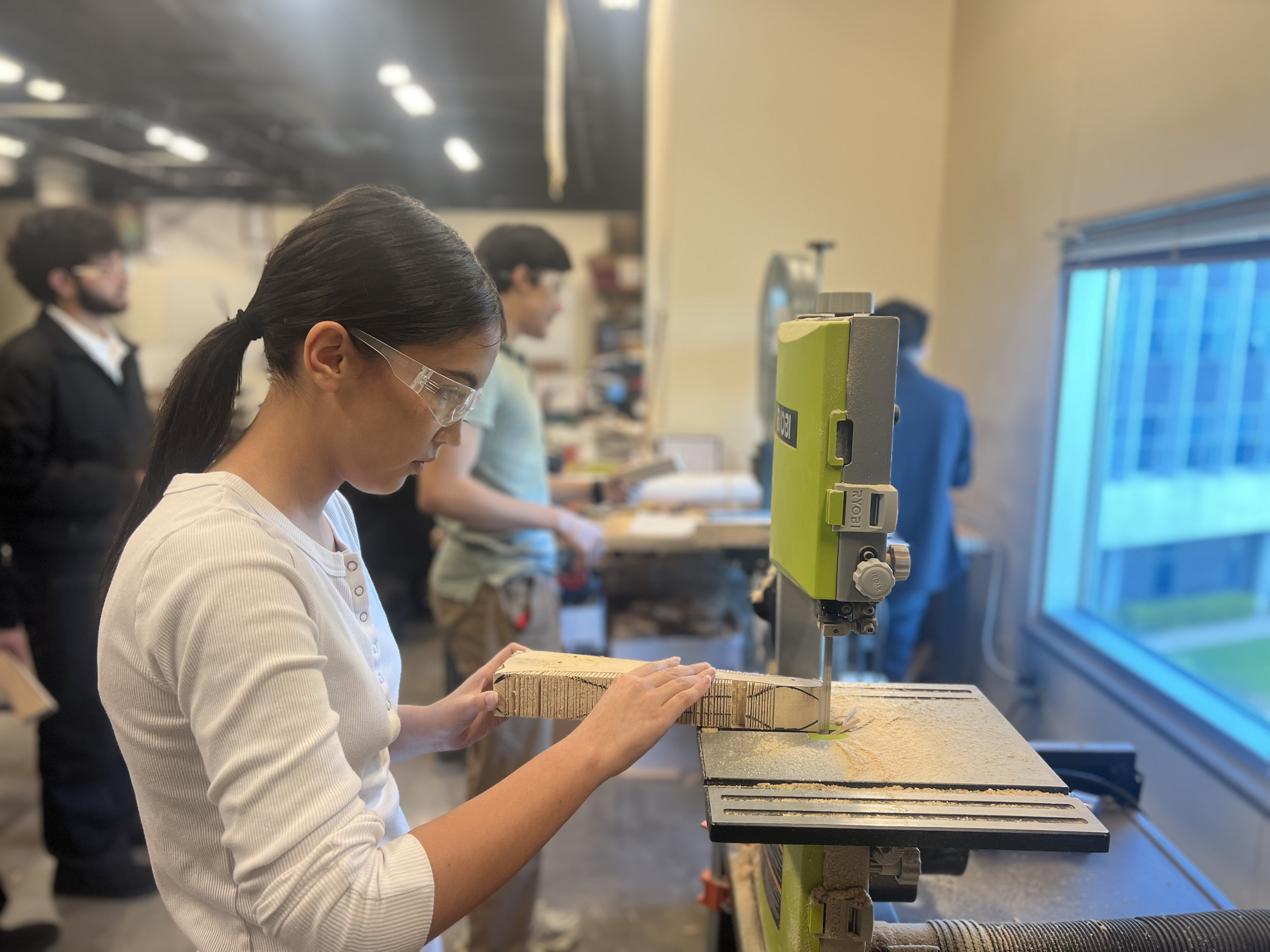 A young woman in a white sweater is operating a power tool at a workbench in what appears to be a workshop or studio setting, with other people visible in the background.