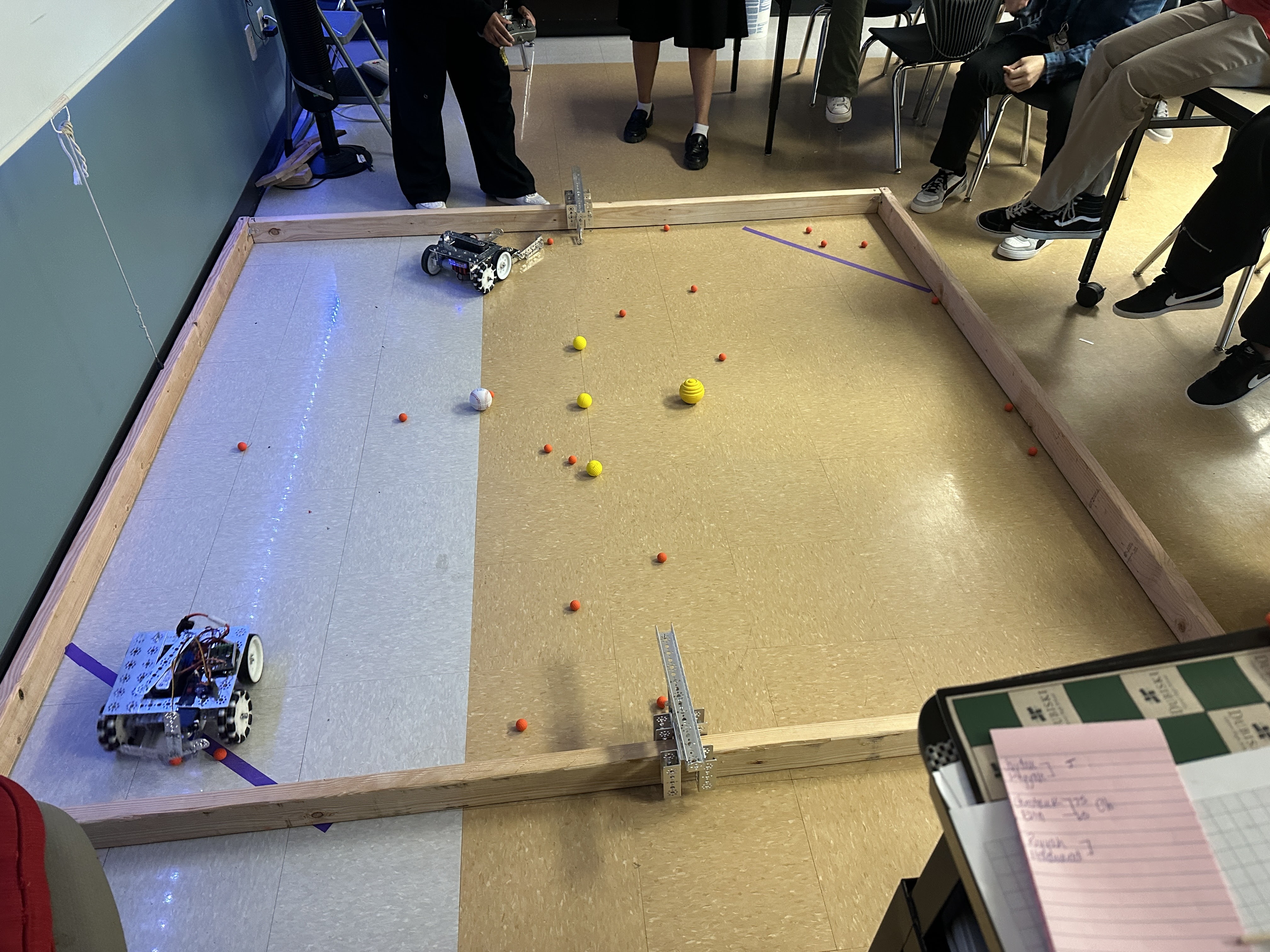 The image shows a large sandbox-like area with various small objects, likely representing a robotics or engineering project, with several people observing or interacting with the setup.
