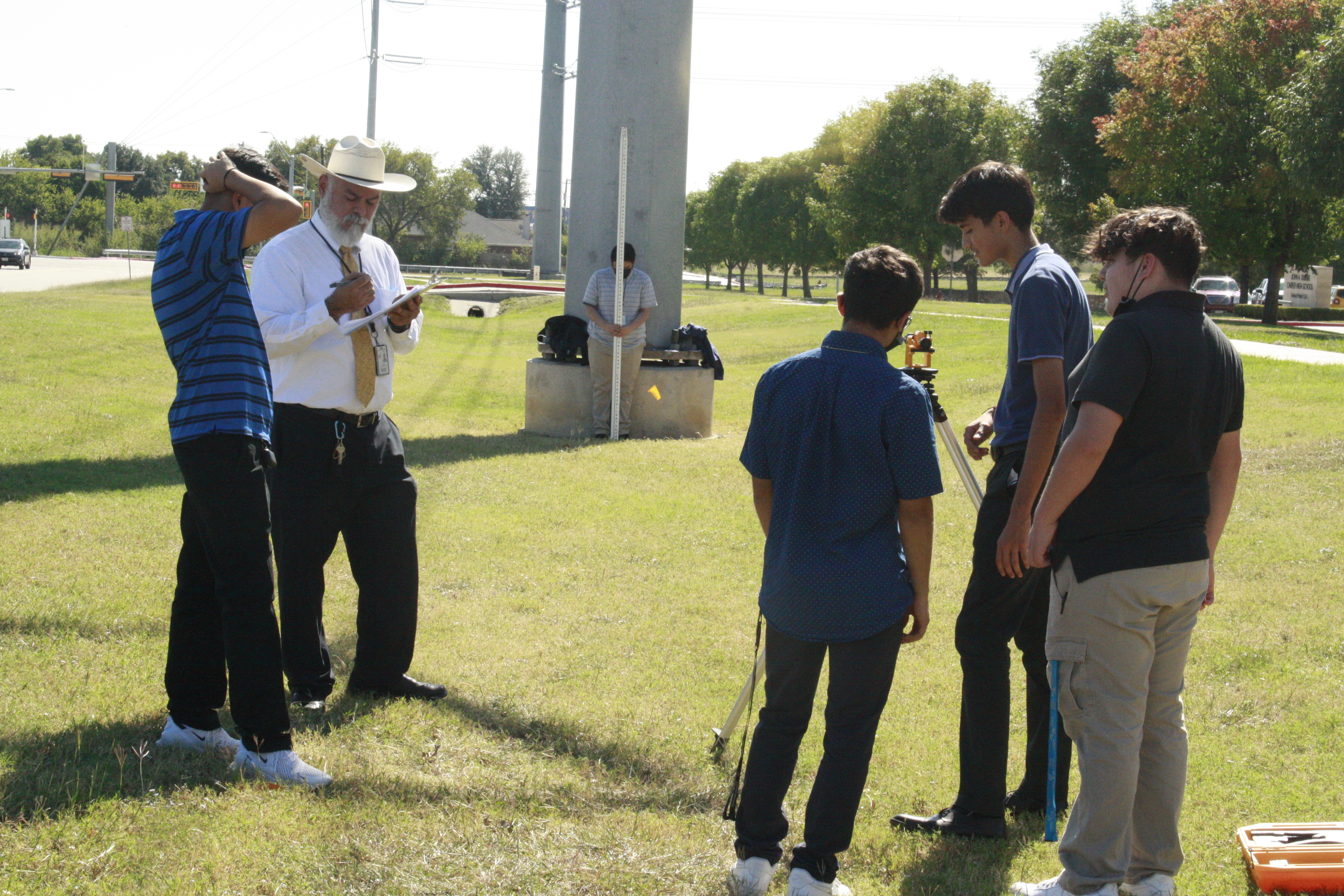 A group of people, some wearing cowboy hats, standing on a grassy field with trees and a flag pole in the background.