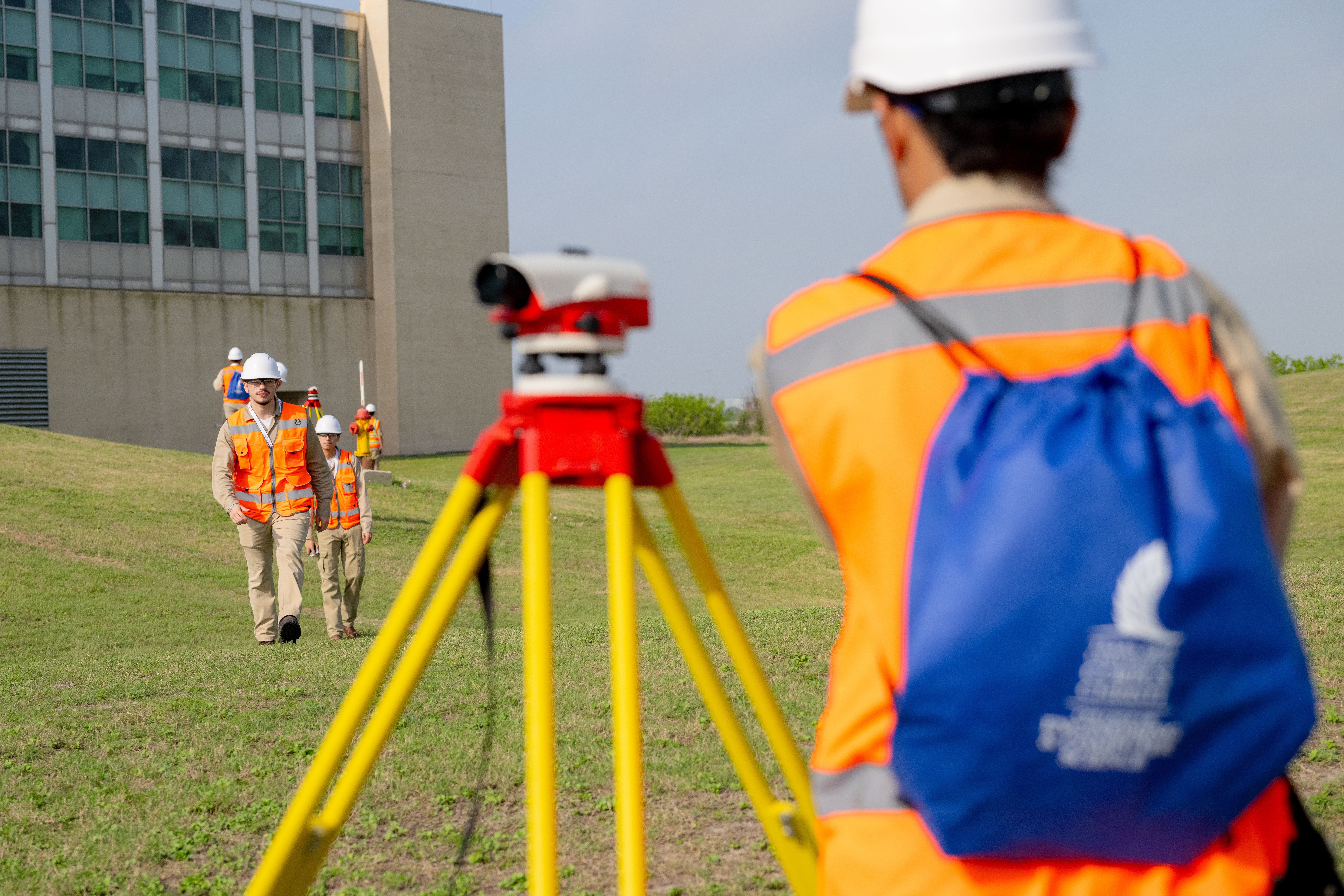 Construction workers in high-visibility vests and hard hats are surveying the site, with a surveying instrument in the foreground and a building in the background.