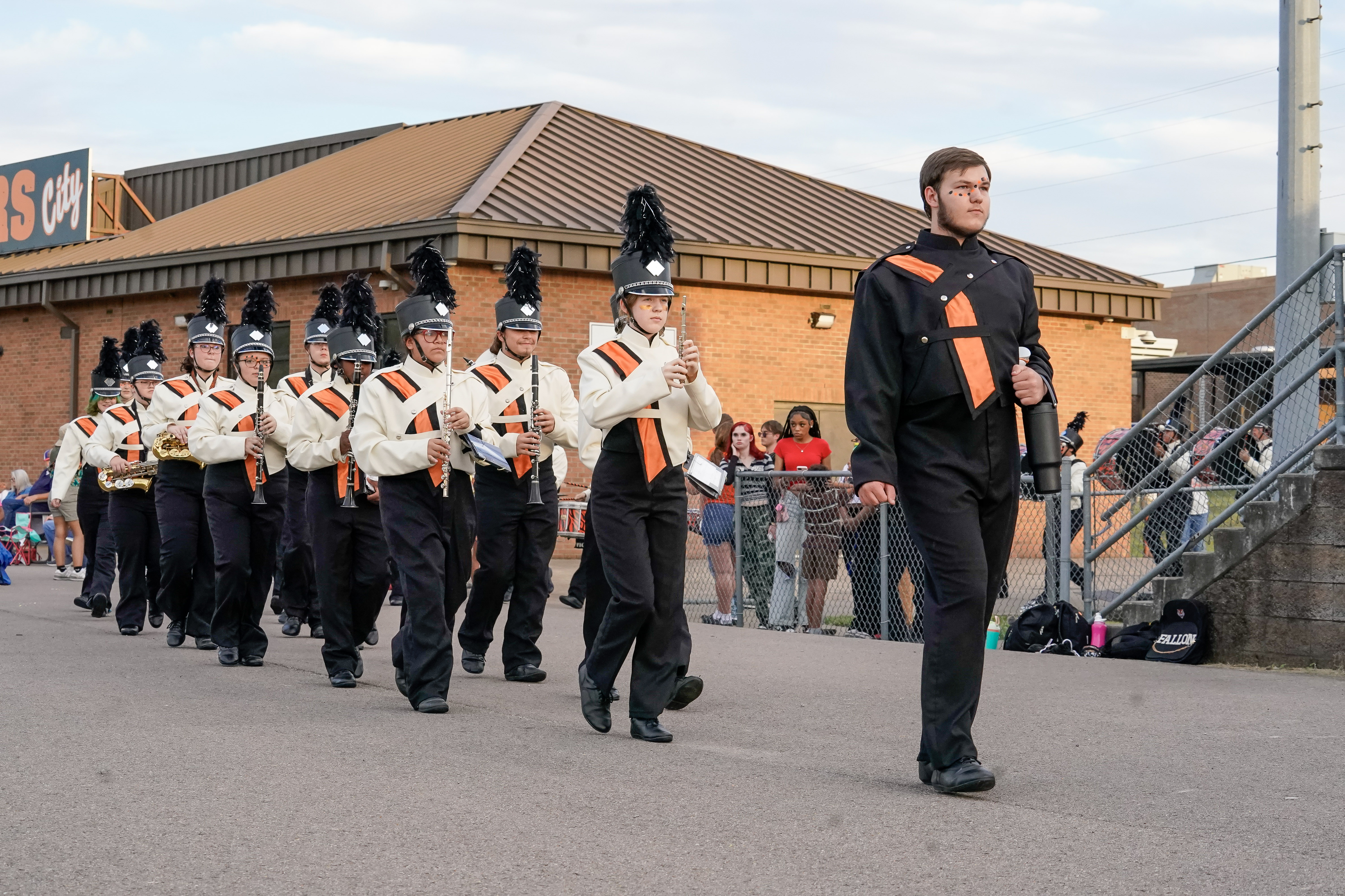 A high school marching band in black, white, and orange uniforms parades outside, led by a drum major. Band members play instruments while spectators watch behind a chain-link fence.