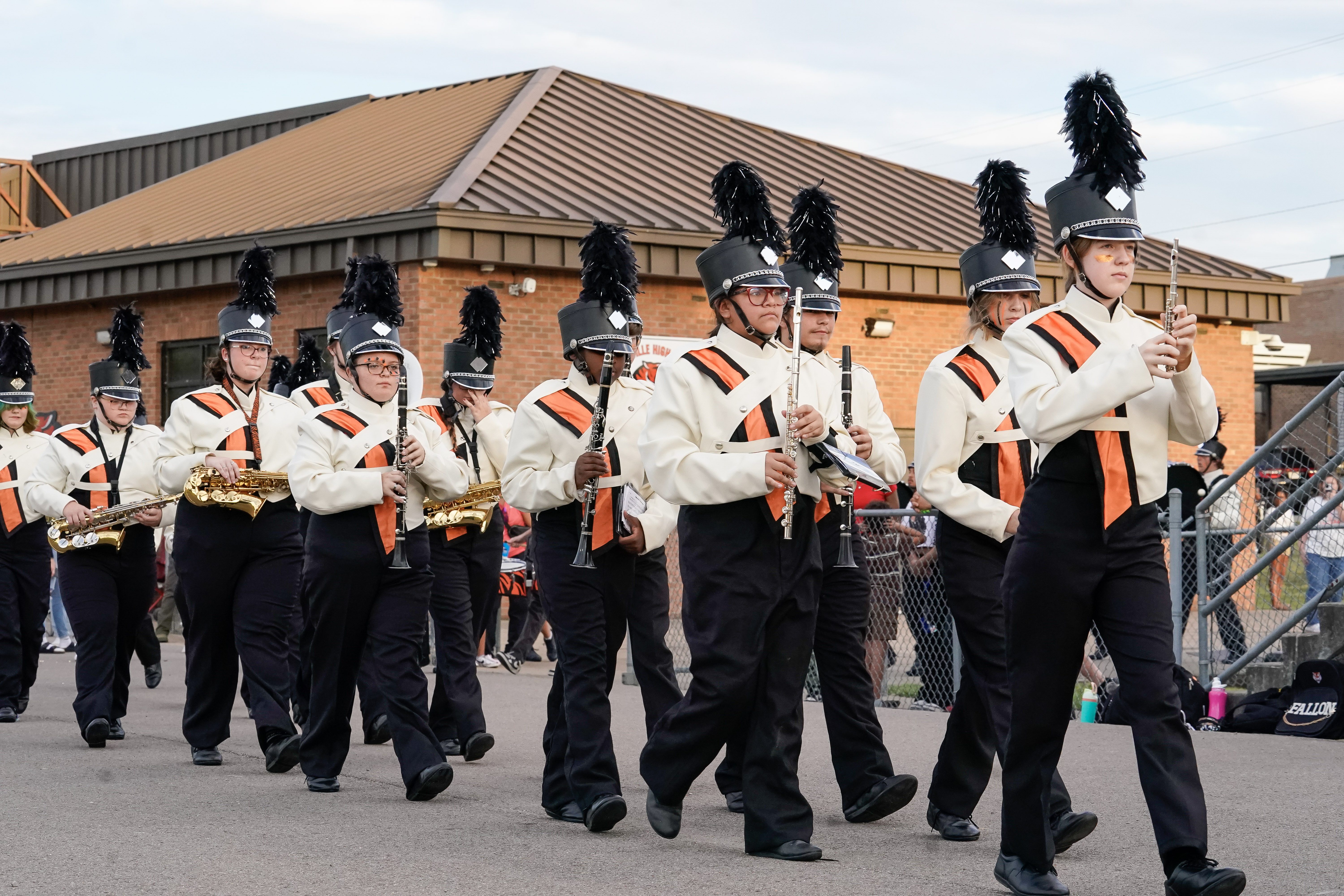 A high school marching band in white and black uniforms with feathered hats plays instruments as they march outside near a brick building and a chain-link fence.