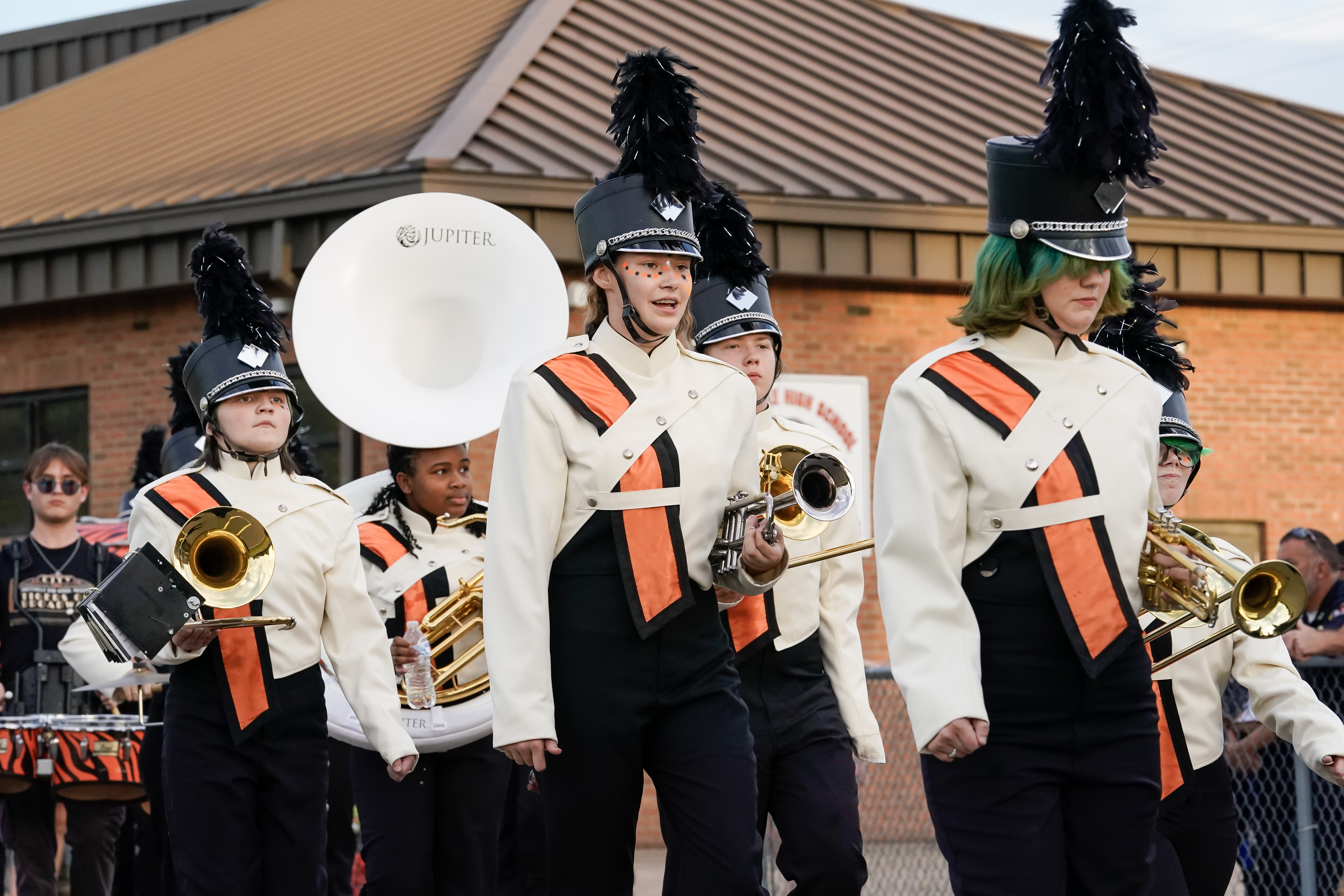 A group of marching band members in white uniforms with black and orange sashes and tall black hats walk in formation, holding brass instruments, with a brick building in the background.