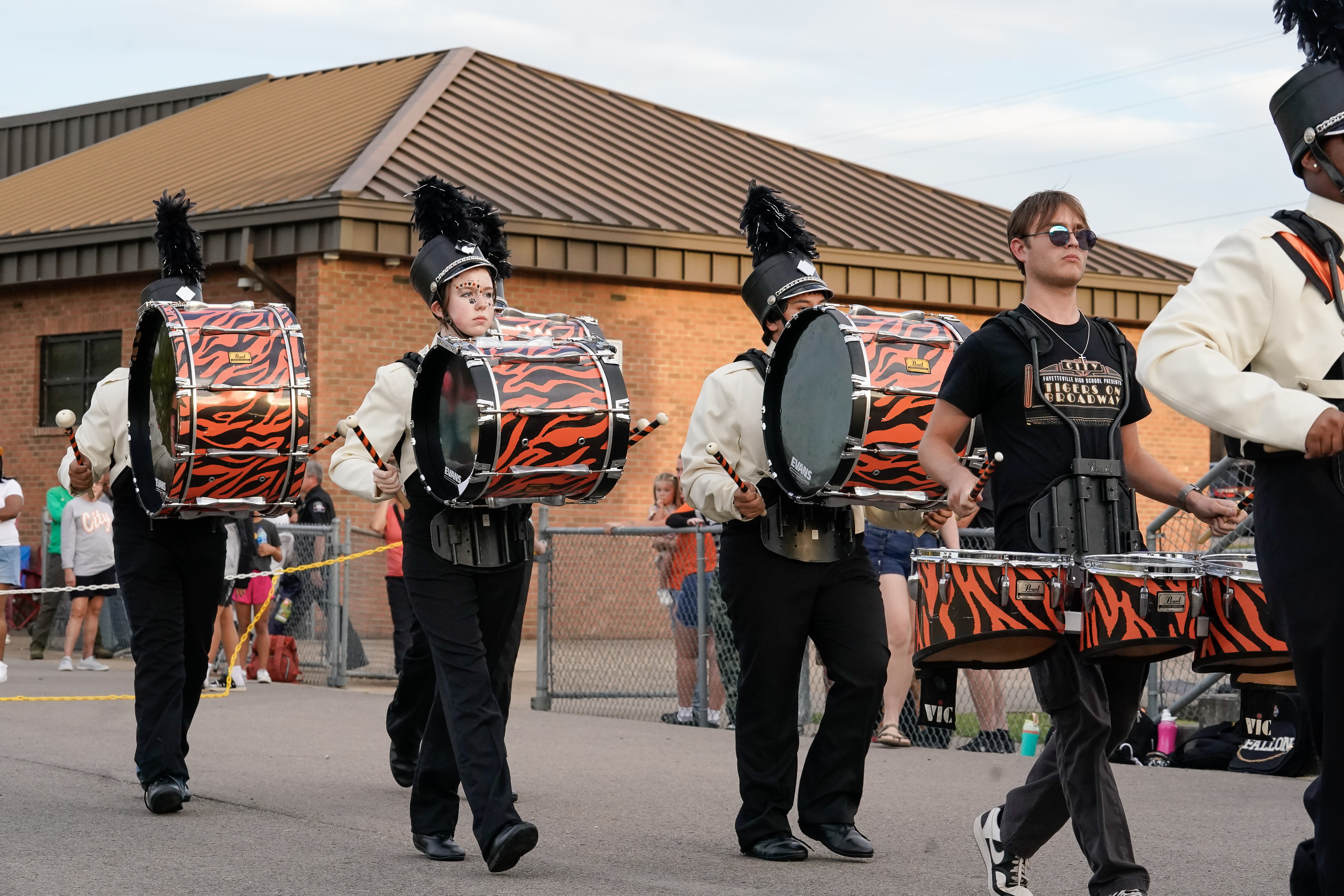 A marching band drumline wearing uniforms and carrying large orange and black tiger-striped bass drums performs outdoors near a crowd and a brick building.