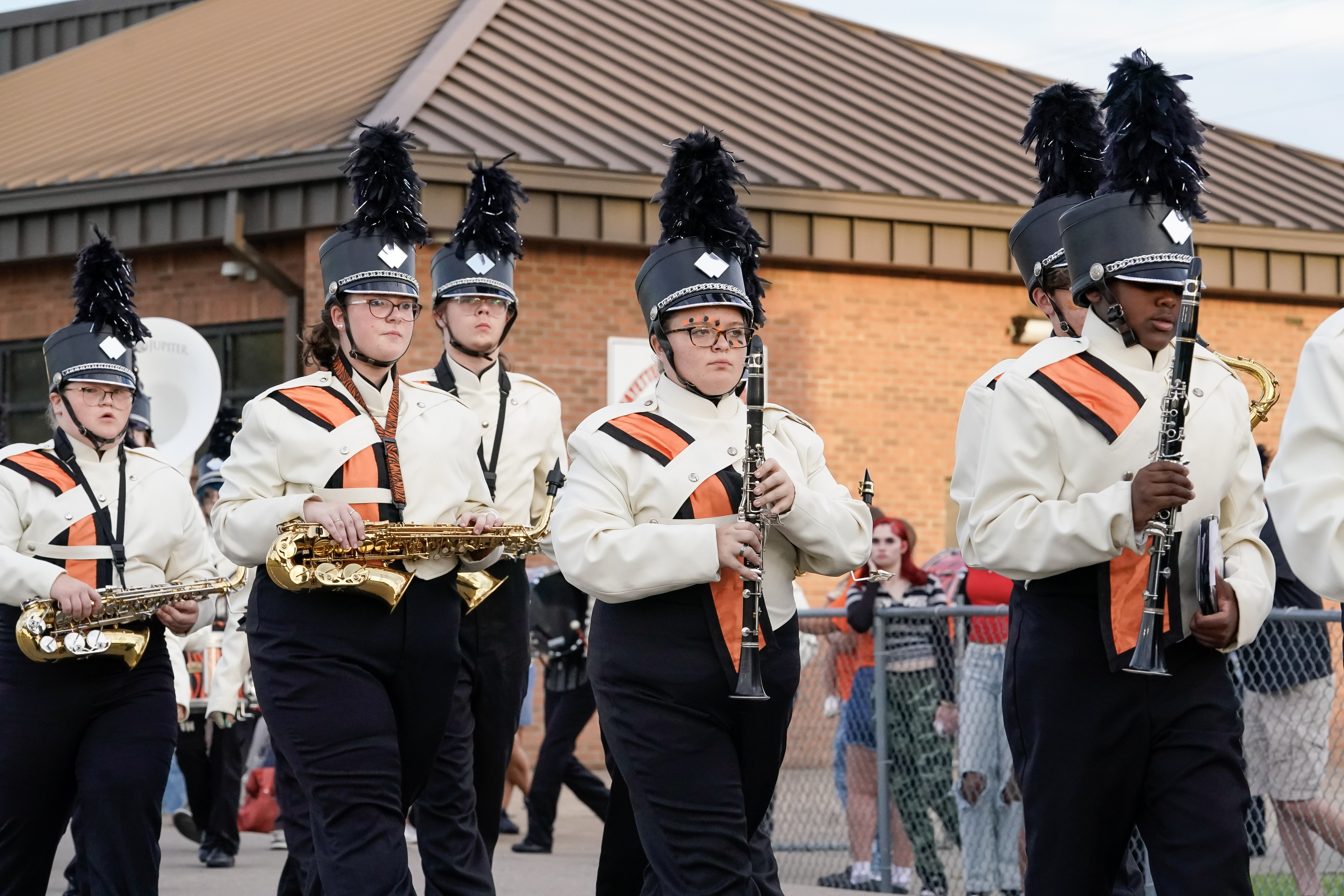 A group of marching band members in white uniforms with black and orange accents walk in formation, playing instruments, including saxophones and clarinets, outside near a brick building.