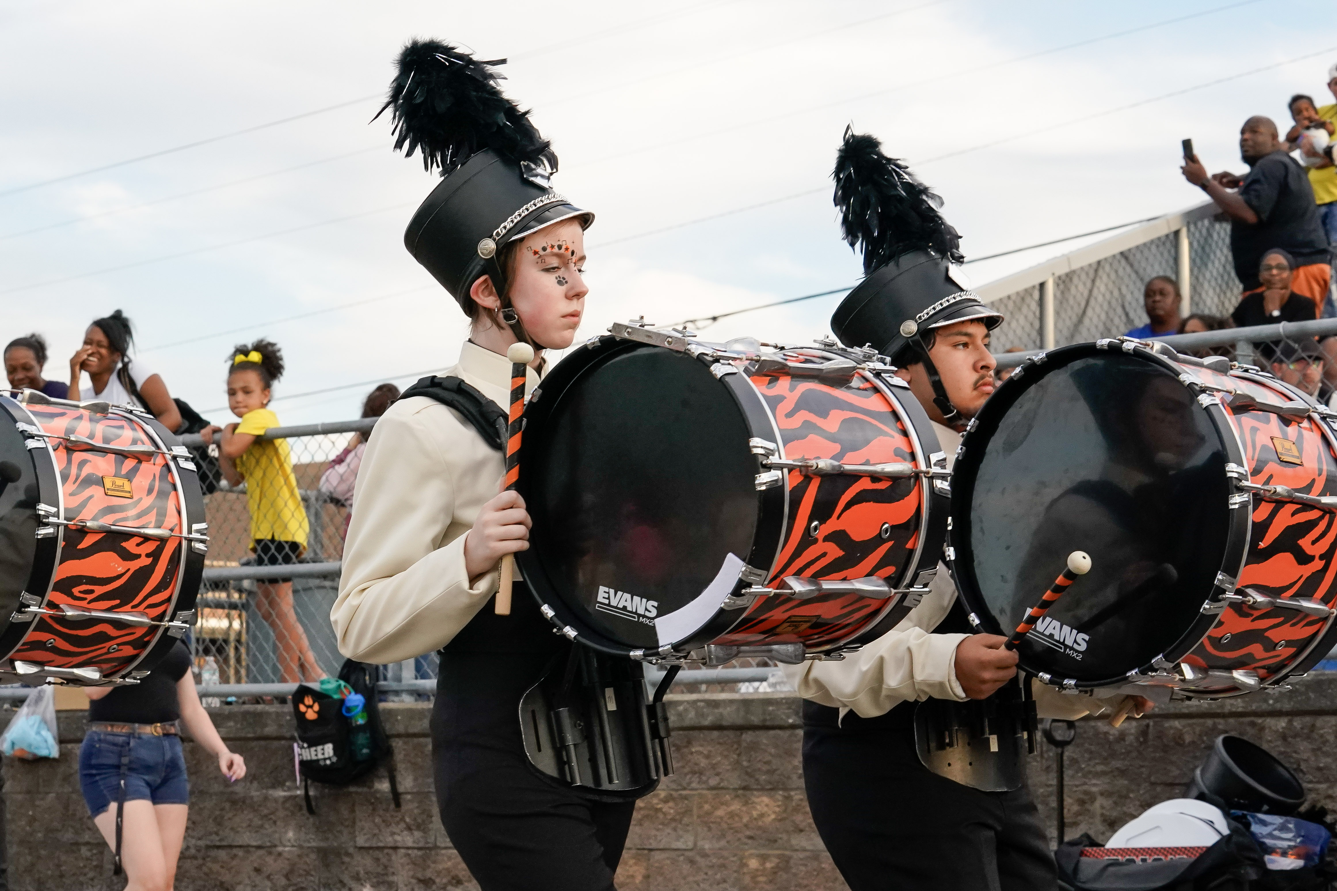 Two marching band members in black hats and uniforms play large orange and black drums during a parade, with spectators watching from behind a fence in the background.