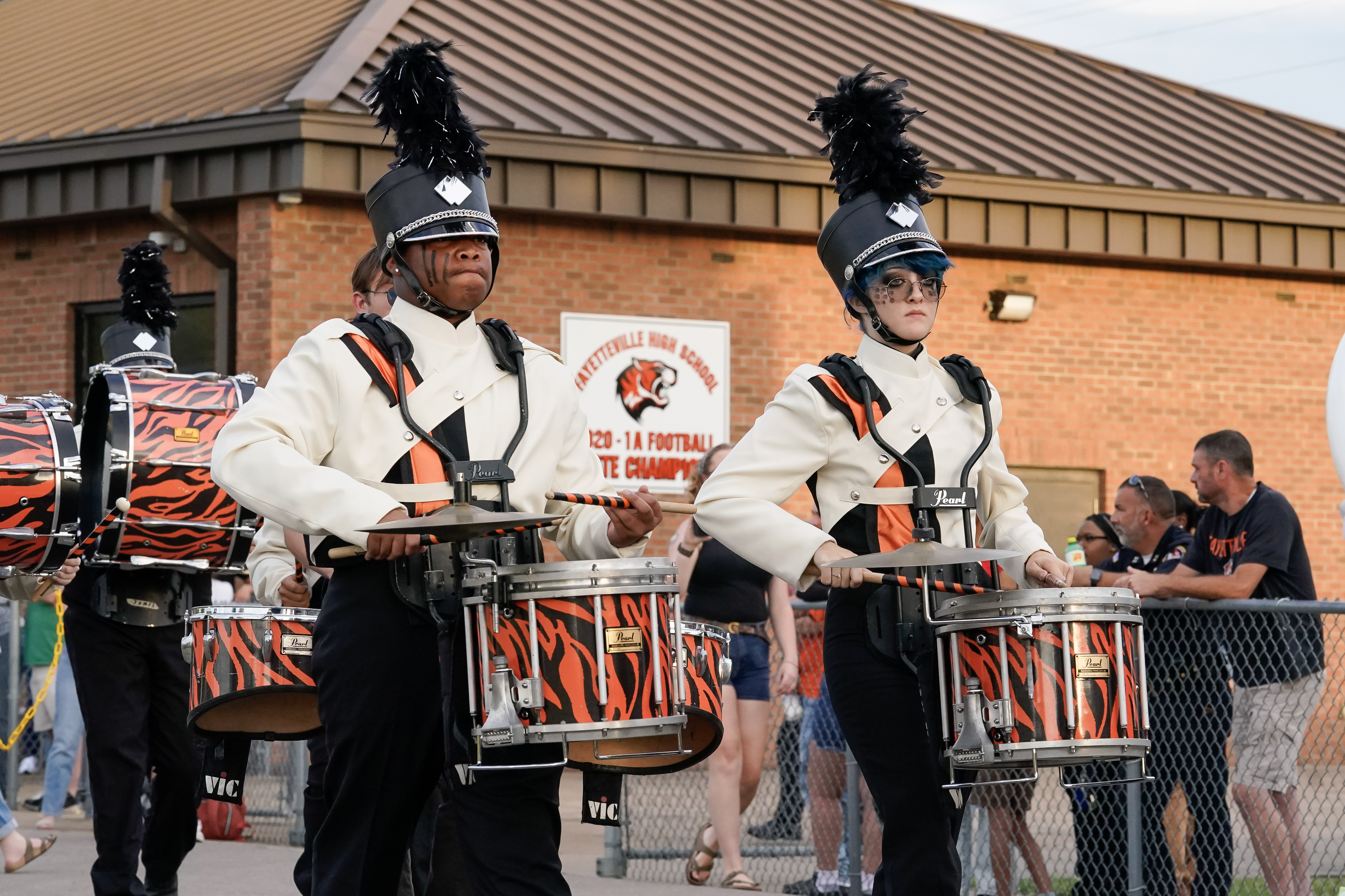 Two marching band drummers in black hats and white uniforms with orange and black drum covers perform in a parade, with spectators and a brick building in the background.