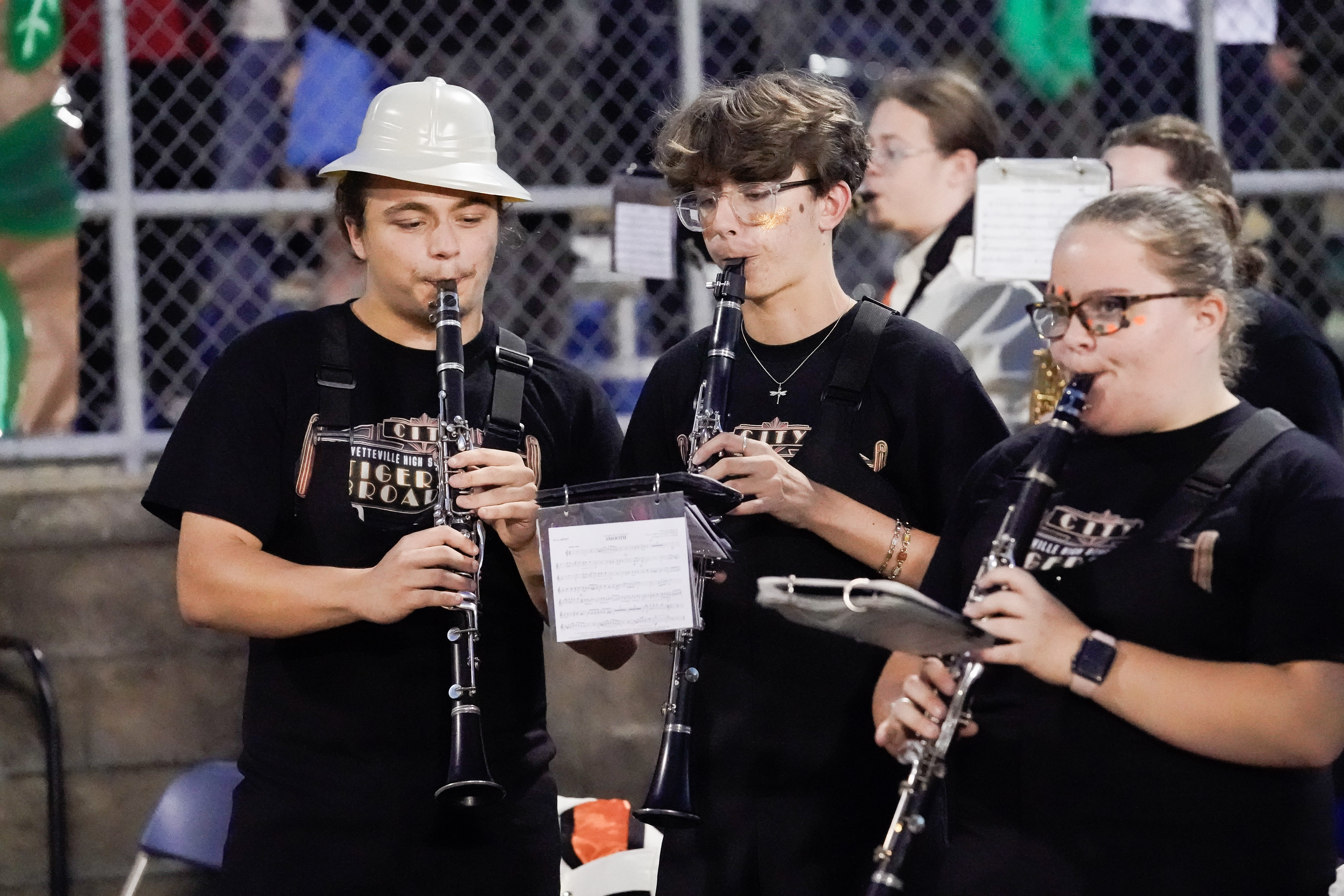 Three marching band members in black shirts play clarinets at an outdoor event. The person on the left wears a white helmet, while the others have glasses. Sheet music is attached to their instruments. A chain-link fence is in the background.
