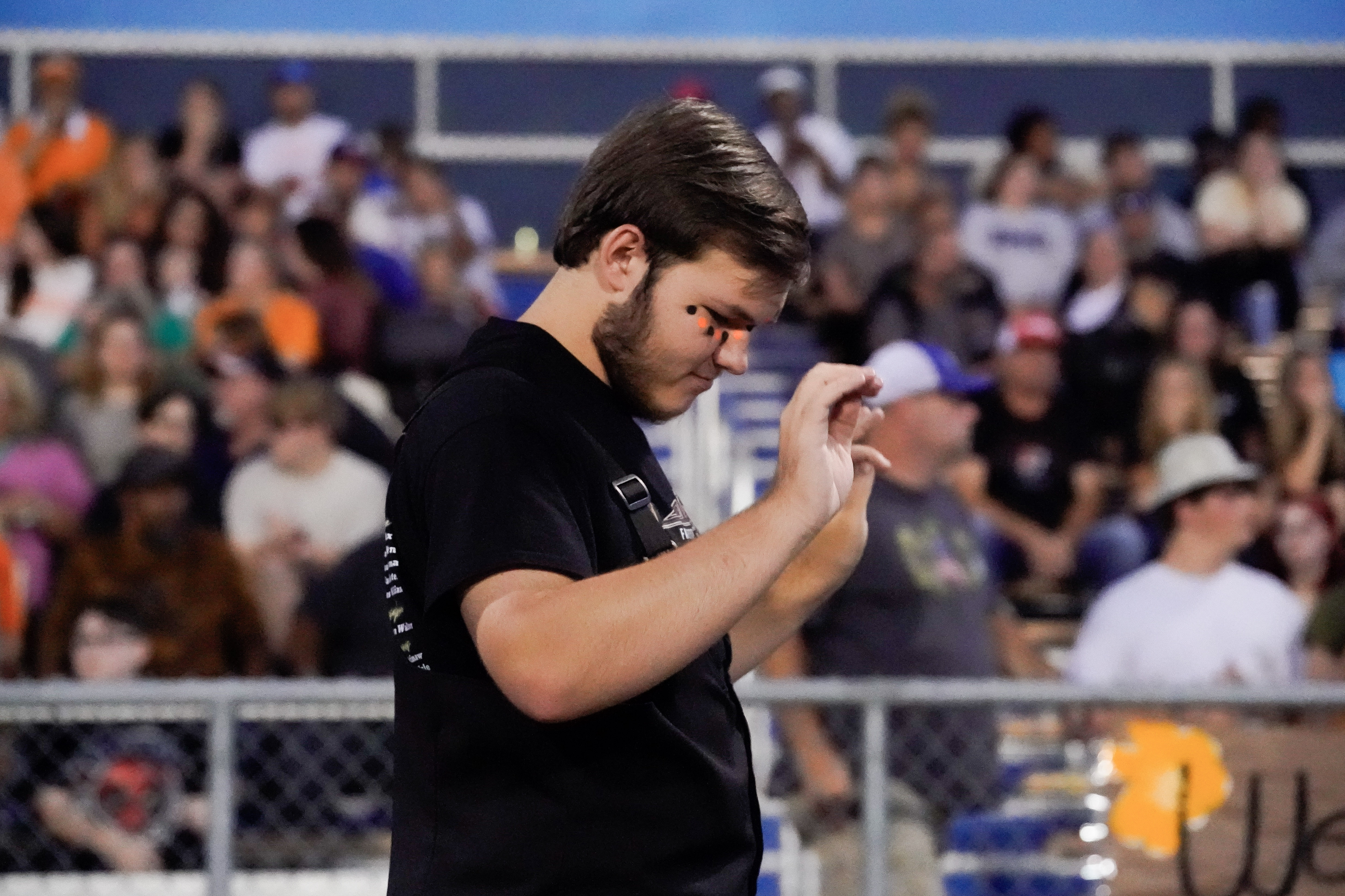 A young man in a black shirt stands with his eyes closed and hands raised, appearing focused, at an outdoor event with blurred spectators in the background.
