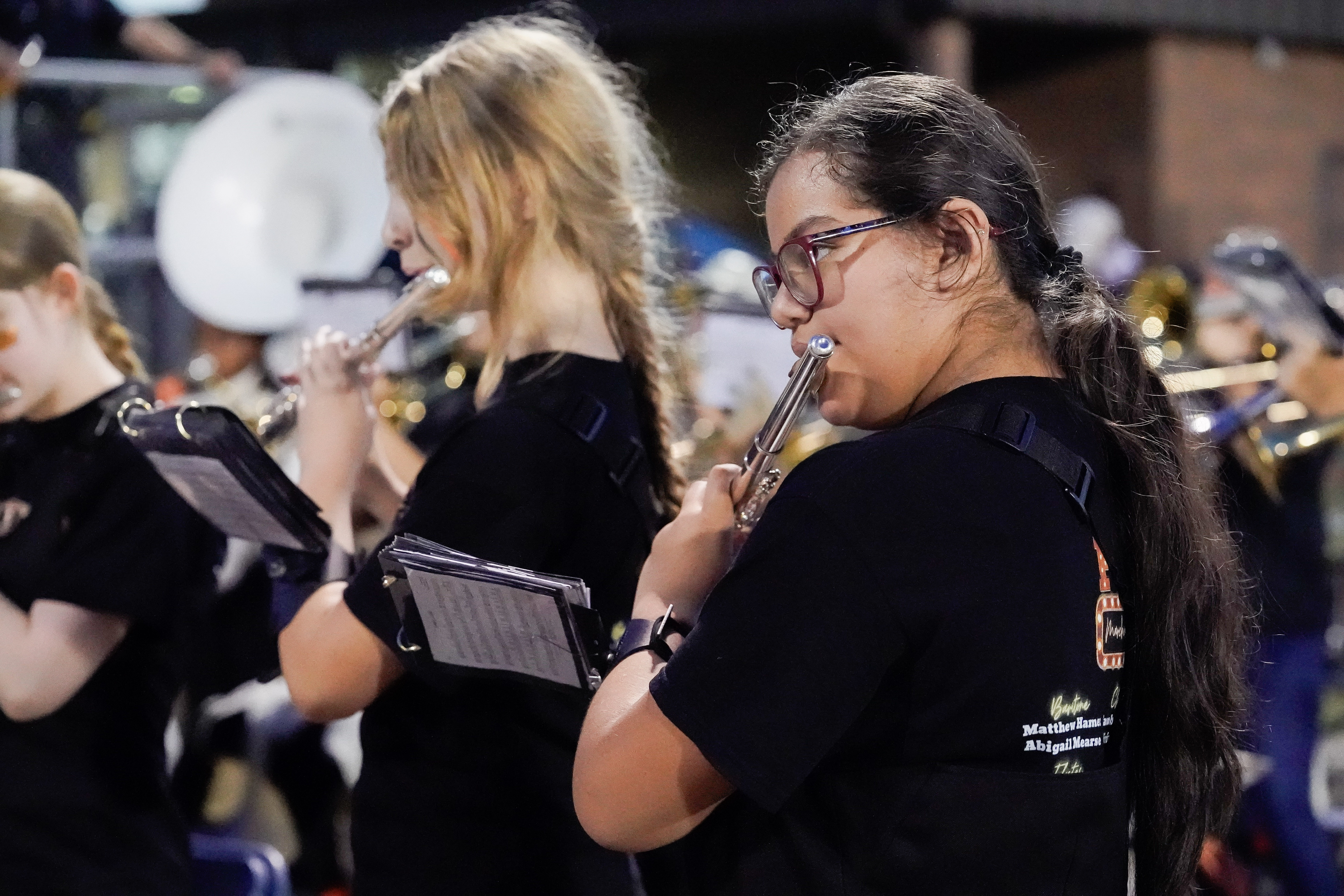 Two girls in black shirts play flutes in a marching band. They hold sheet music and focus on their performance, surrounded by other band members with brass instruments in the background.