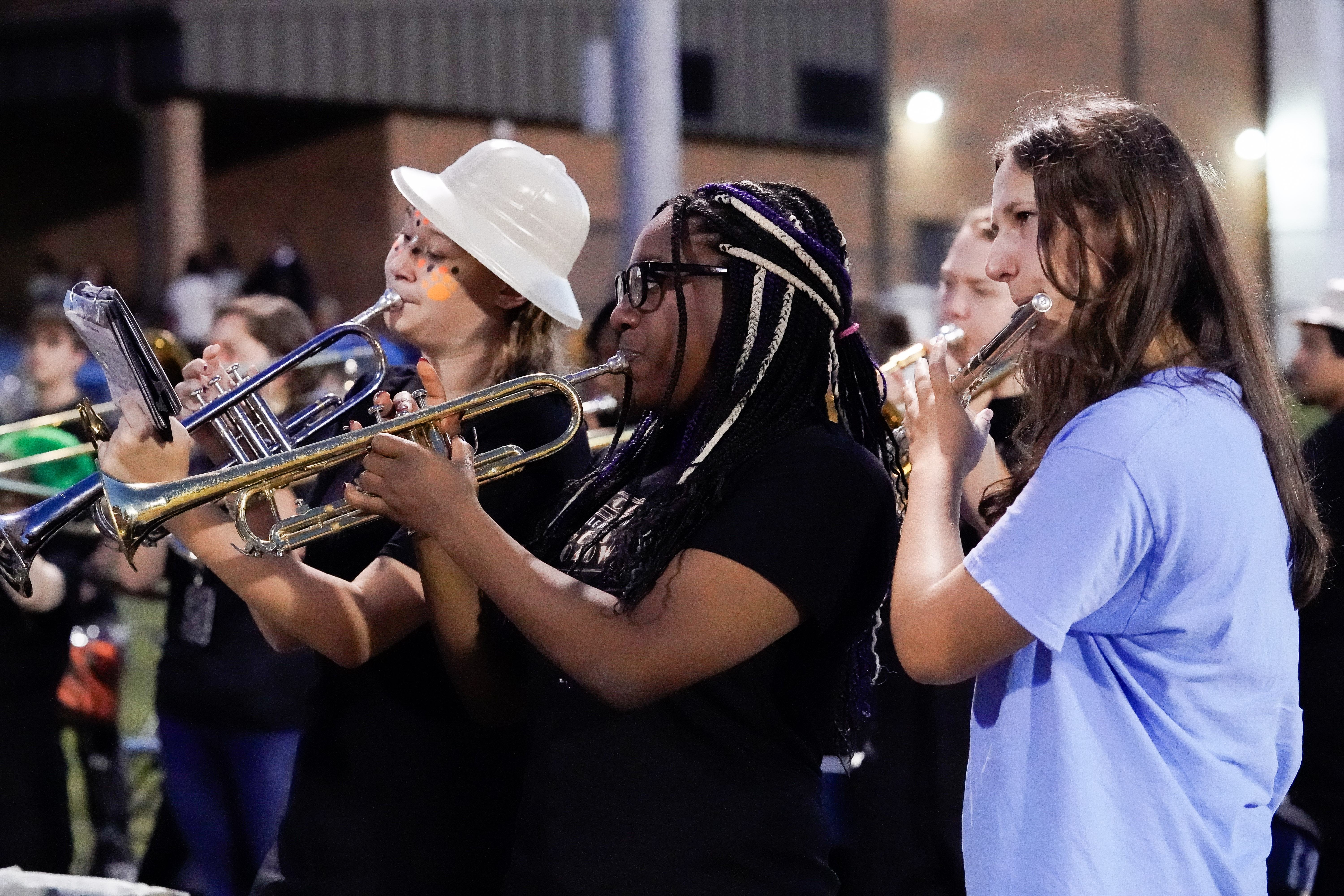 Three students play trumpets and a flute outdoors at night. One wears a white helmet and face paint, another has braids and glasses, and the third wears a blue shirt. People and buildings are visible in the background.