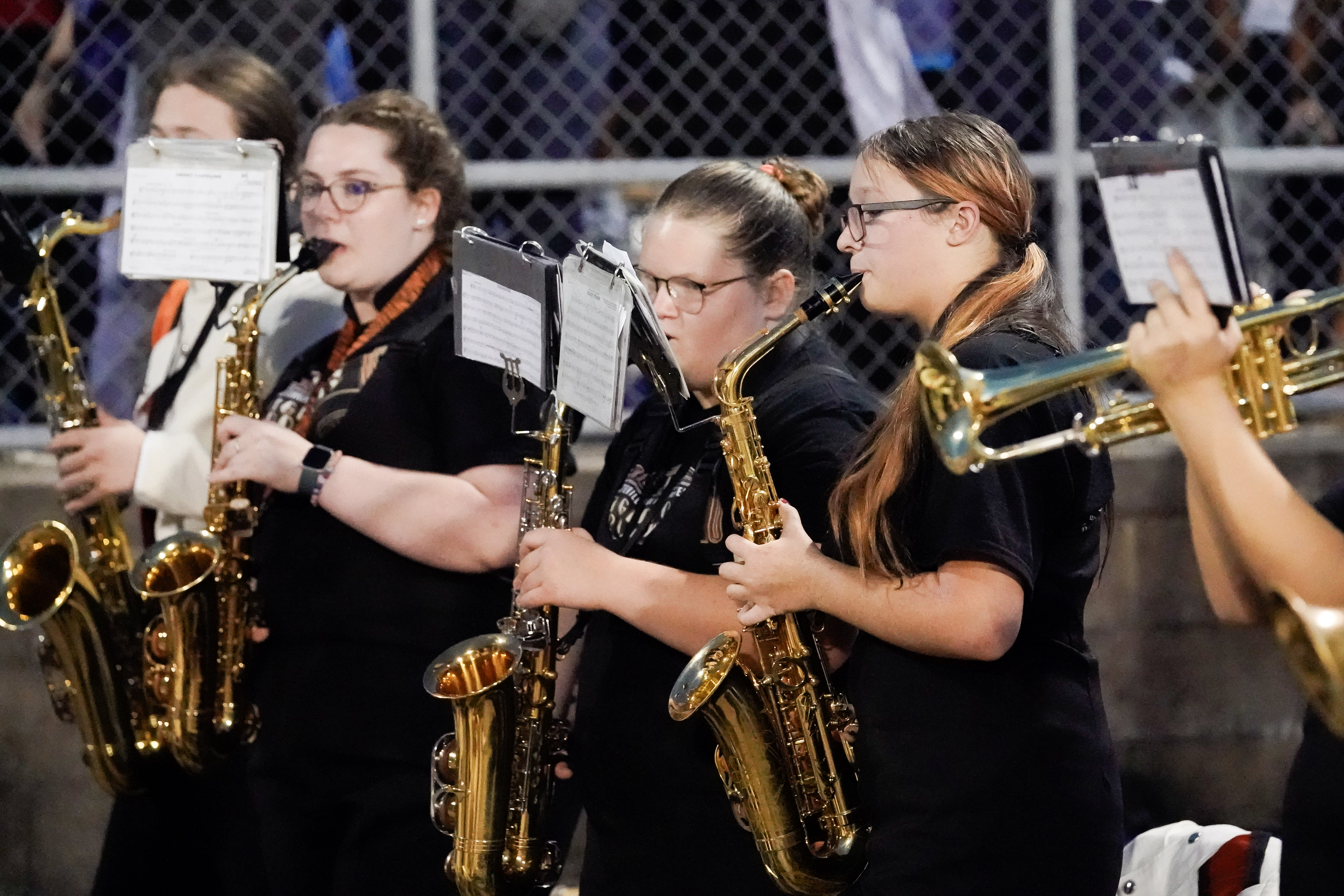 Three band members wearing black shirts play saxophones at an outdoor event. Music sheets are attached to their instruments, and a partially visible trumpet is in the foreground. A chain-link fence is in the background.