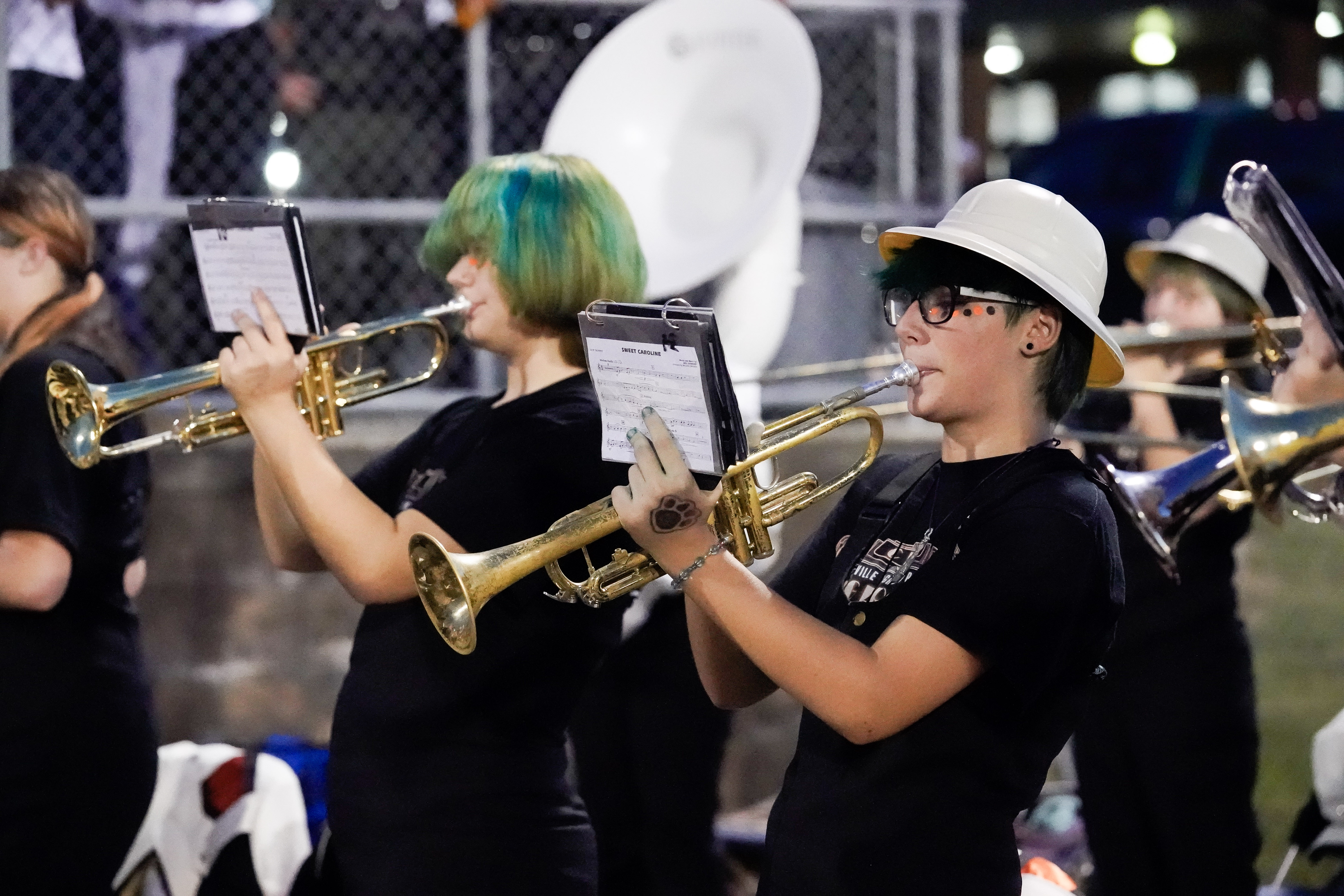Two band members play trumpets during an outdoor performance at night. Both wear black shirts; one has green hair, and the other wears a white hat and glasses. A sousaphone is visible in the background.