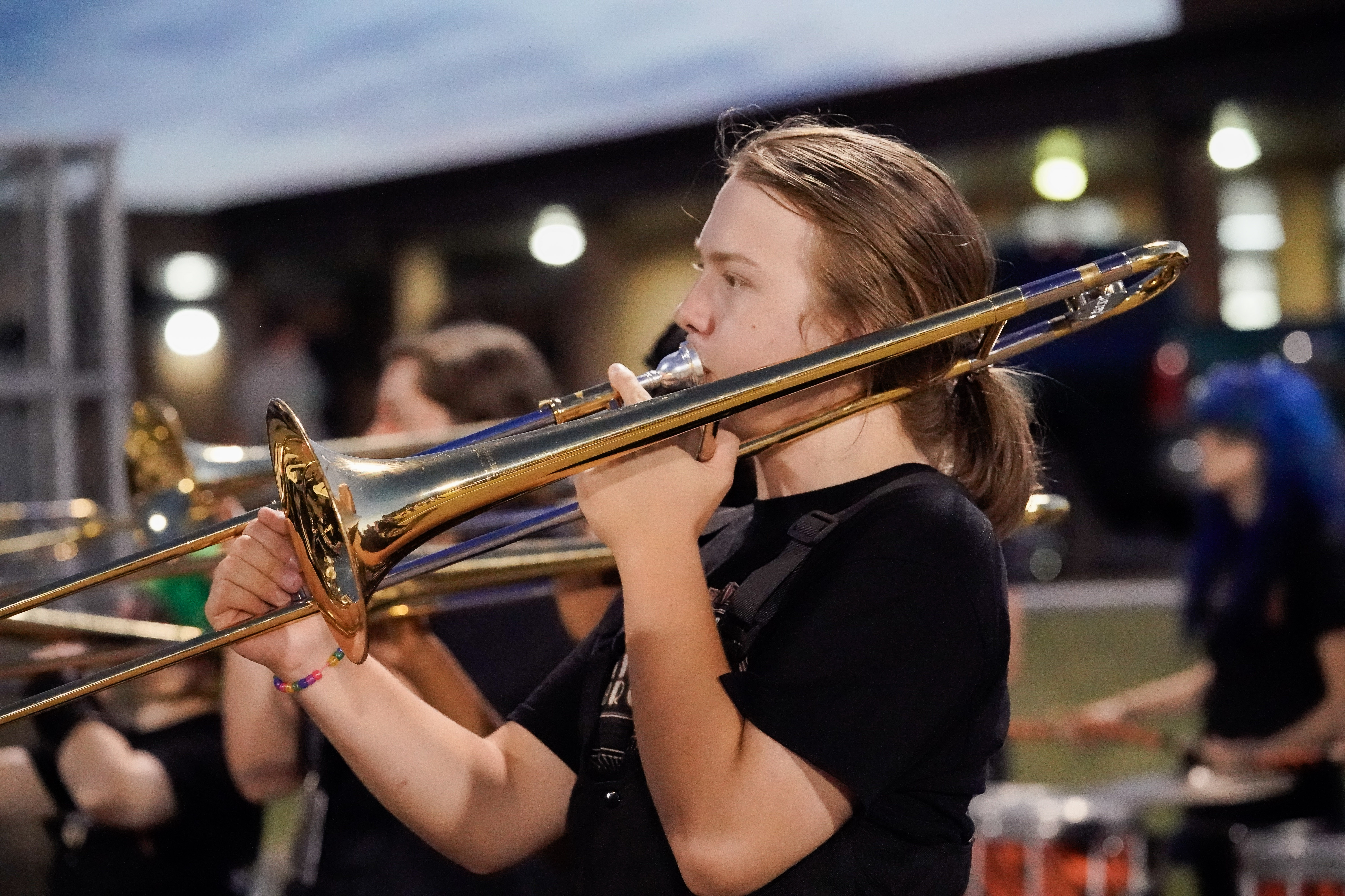 A young person with long hair plays a trombone during an outdoor band performance at dusk, with blurred bandmates and building lights in the background.
