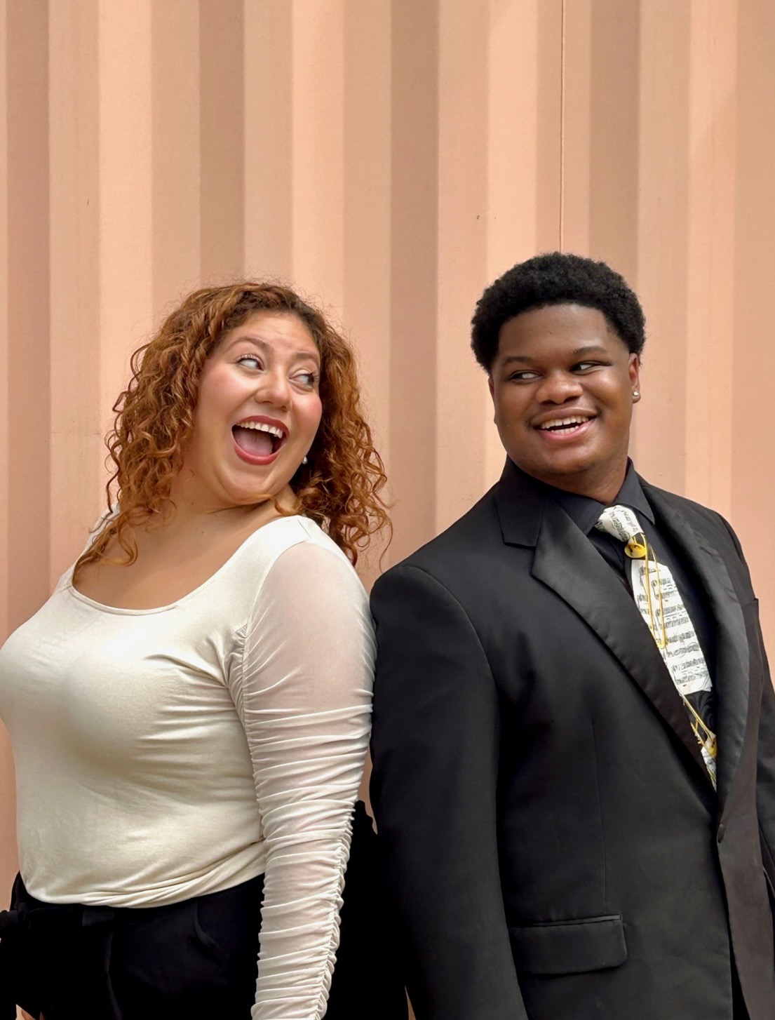 The image shows a smiling woman with curly hair standing next to a smiling man in a suit against a striped background.
