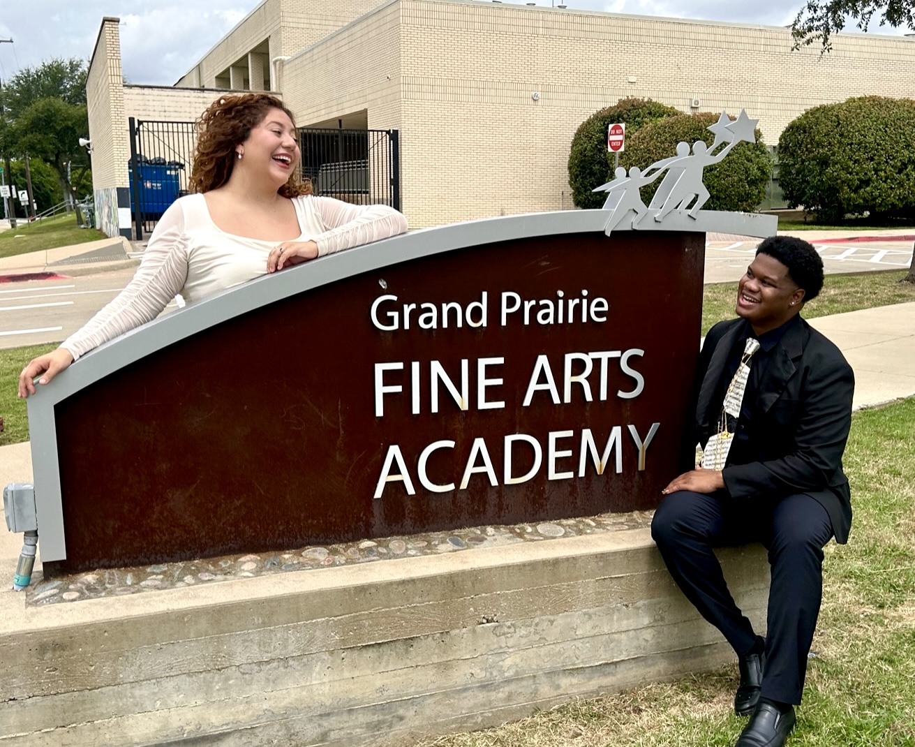 The image shows two people, a woman and a man, posing in front of a sign for the Grand Prairie Fine Arts Academy.