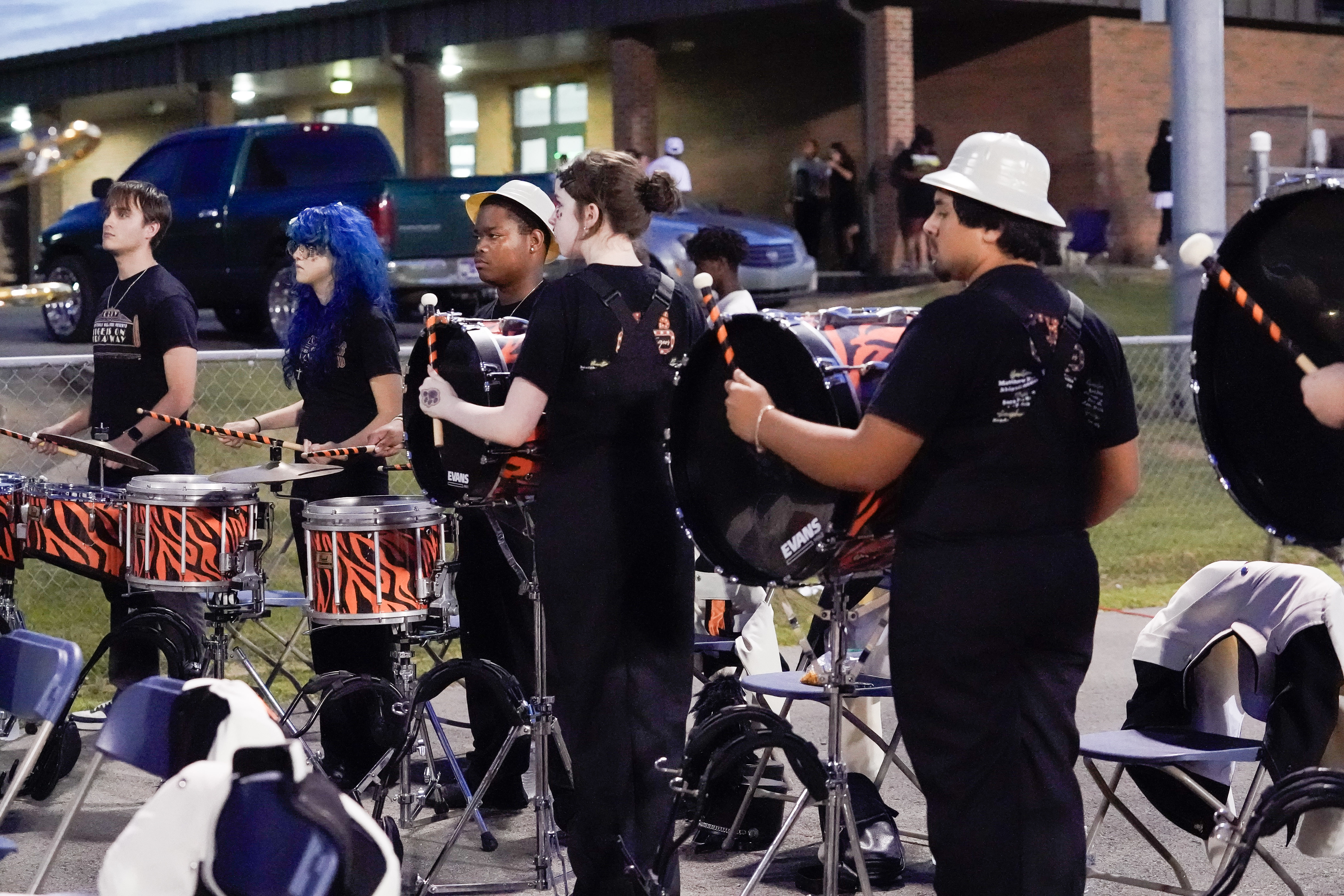 A group of high school students in black uniforms play drums outdoors near a school building at dusk, with some chairs and band equipment around them.