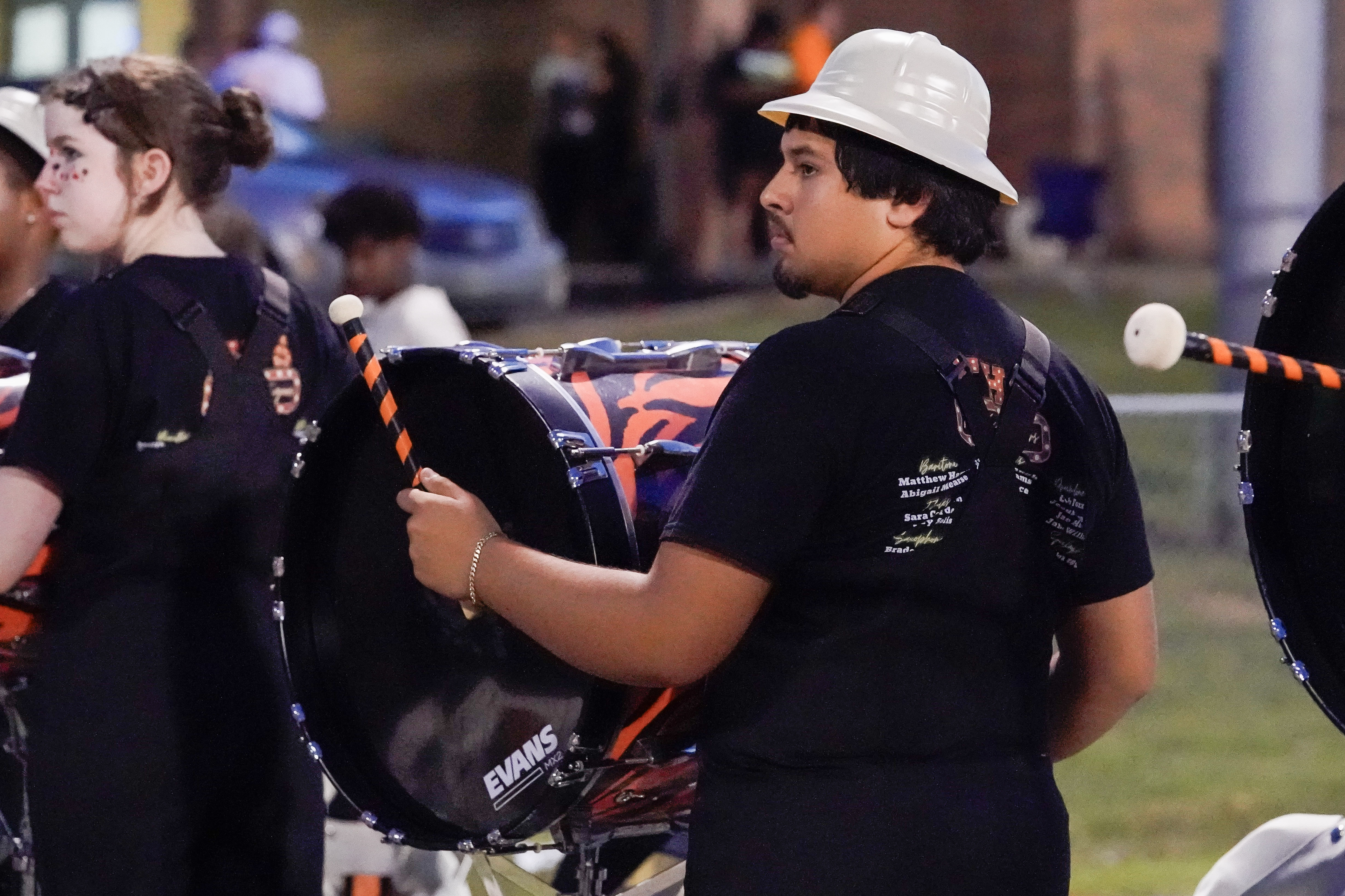 A young man wearing a white hardhat plays a bass drum with a mallet during an outdoor performance. He is part of a drumline and stands beside others in matching black shirts.