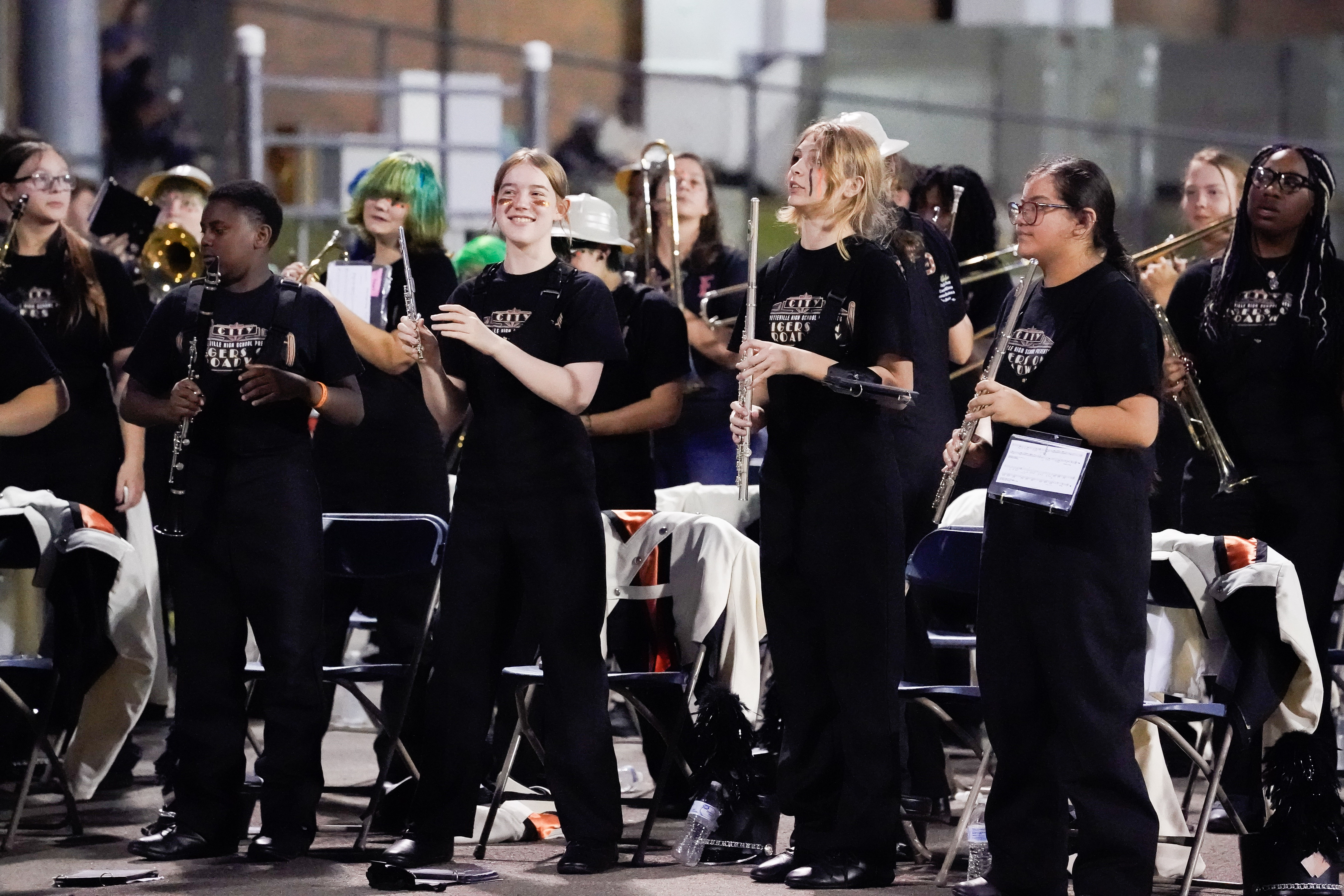 A group of teenagers in matching black uniforms stand and smile while holding musical instruments at an outdoor event, with empty chairs and scattered jackets around them.