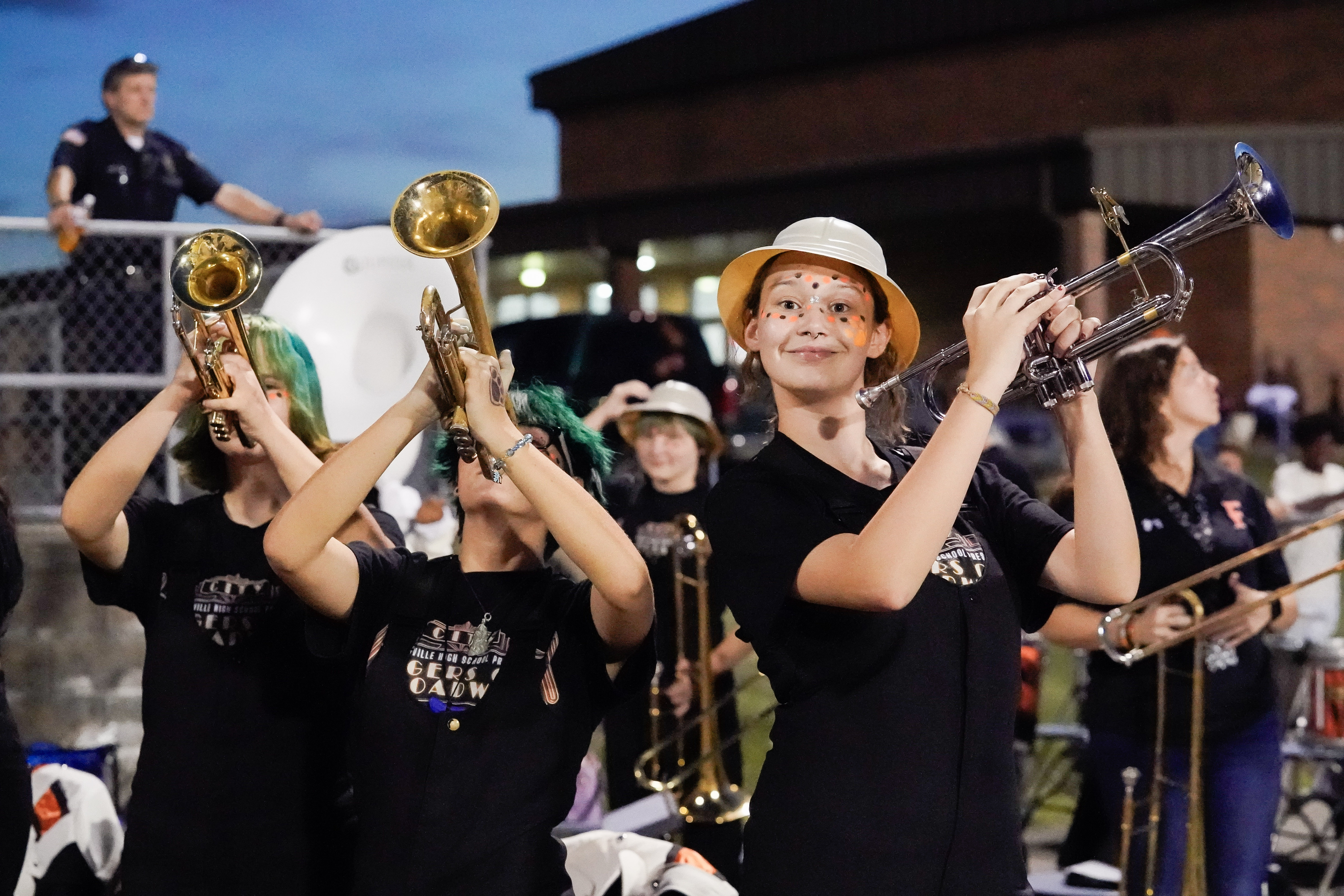 Three young trumpet players in matching black shirts perform outdoors at dusk. One wears a white hat and face paint, smiling at the camera, while the others play with their instruments raised. Spectators and a building are in the background.