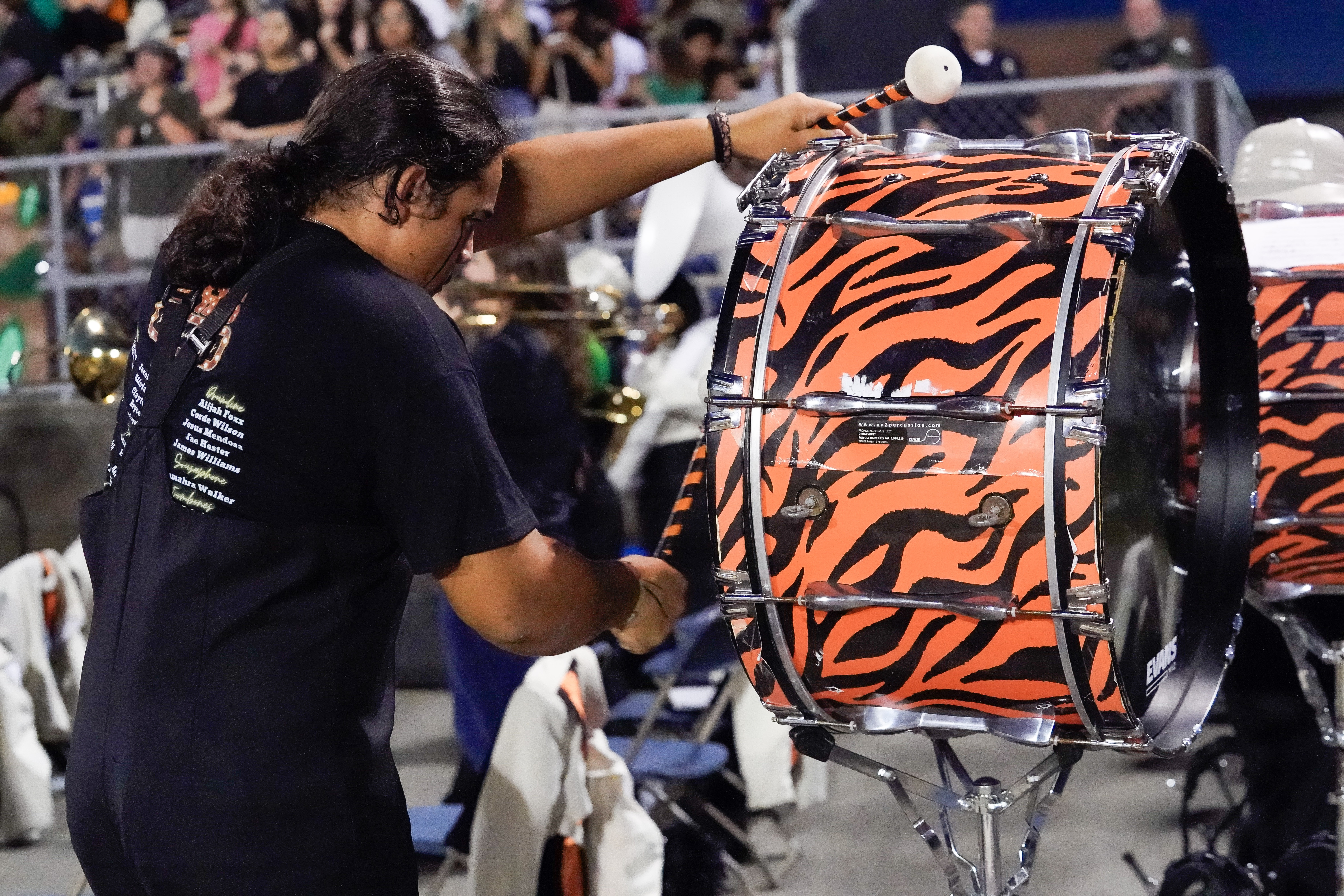 A person wearing black plays a large bass drum with an orange and black tiger stripe pattern at an outdoor event, with a crowd and other musicians visible in the background.