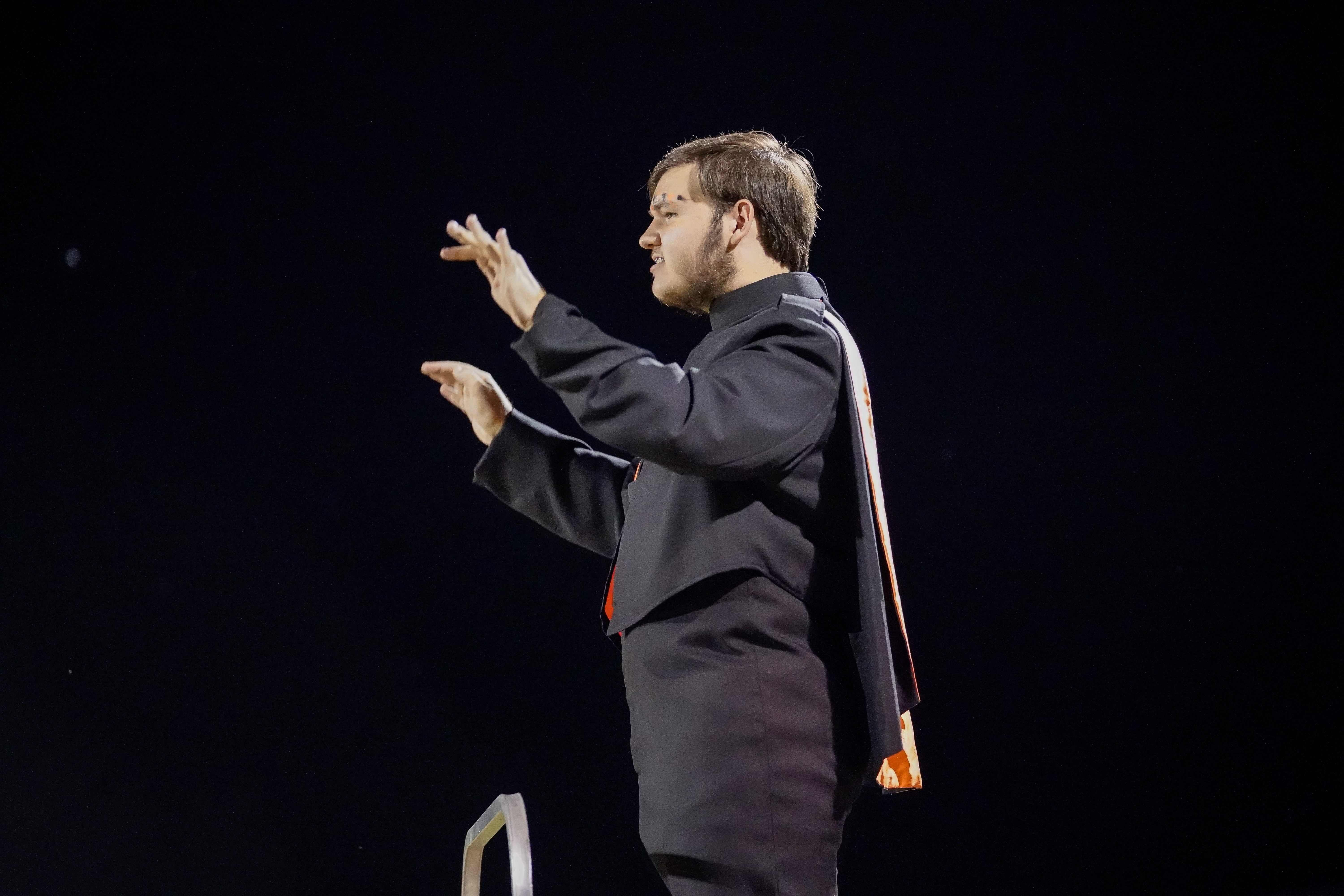A person in a band uniform stands on a platform, facing left, conducting with both hands raised against a dark night sky.