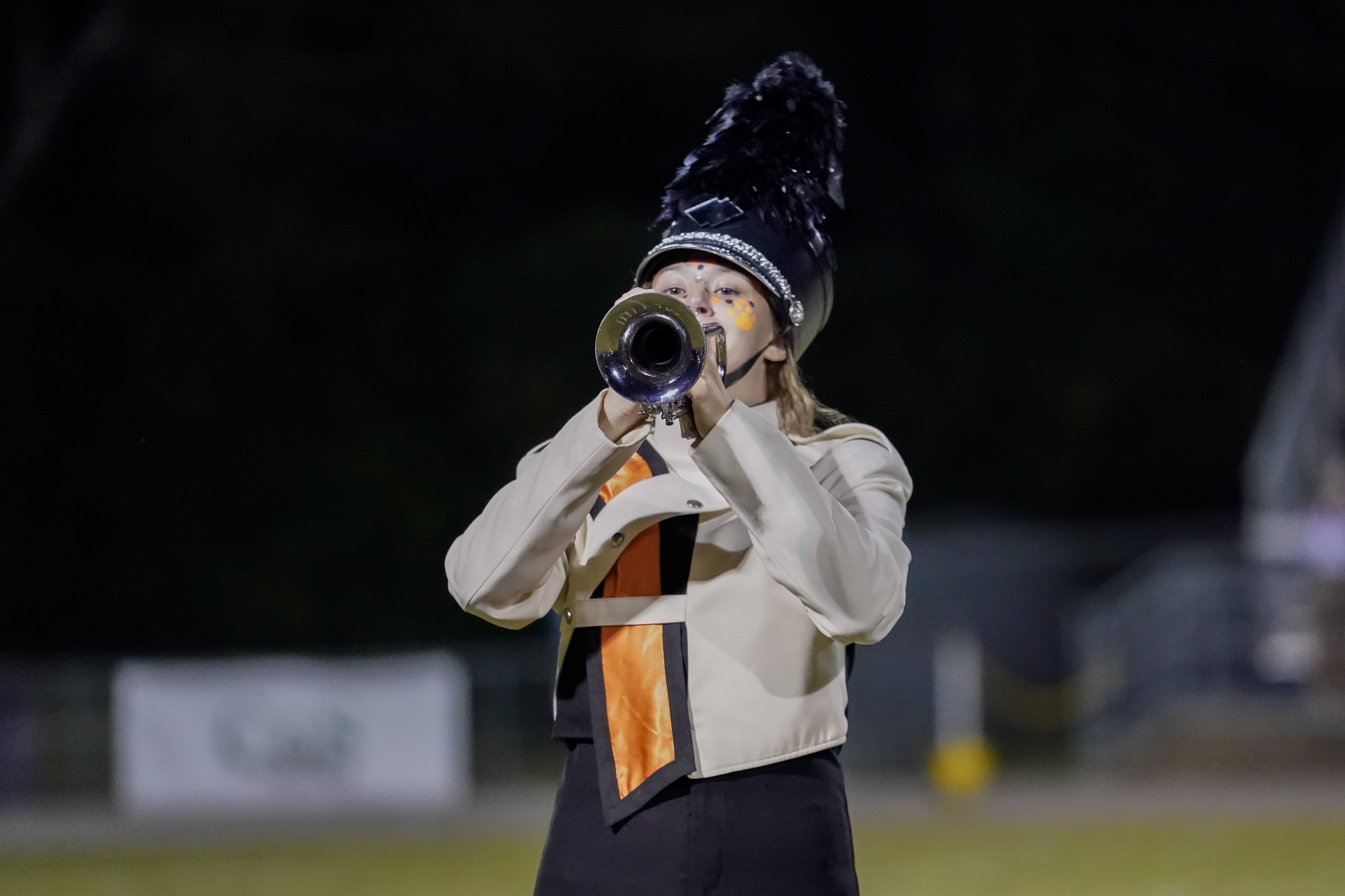 A marching band member in a white and black uniform with an orange sash plays a trumpet outdoors at night, wearing a tall feathered hat.