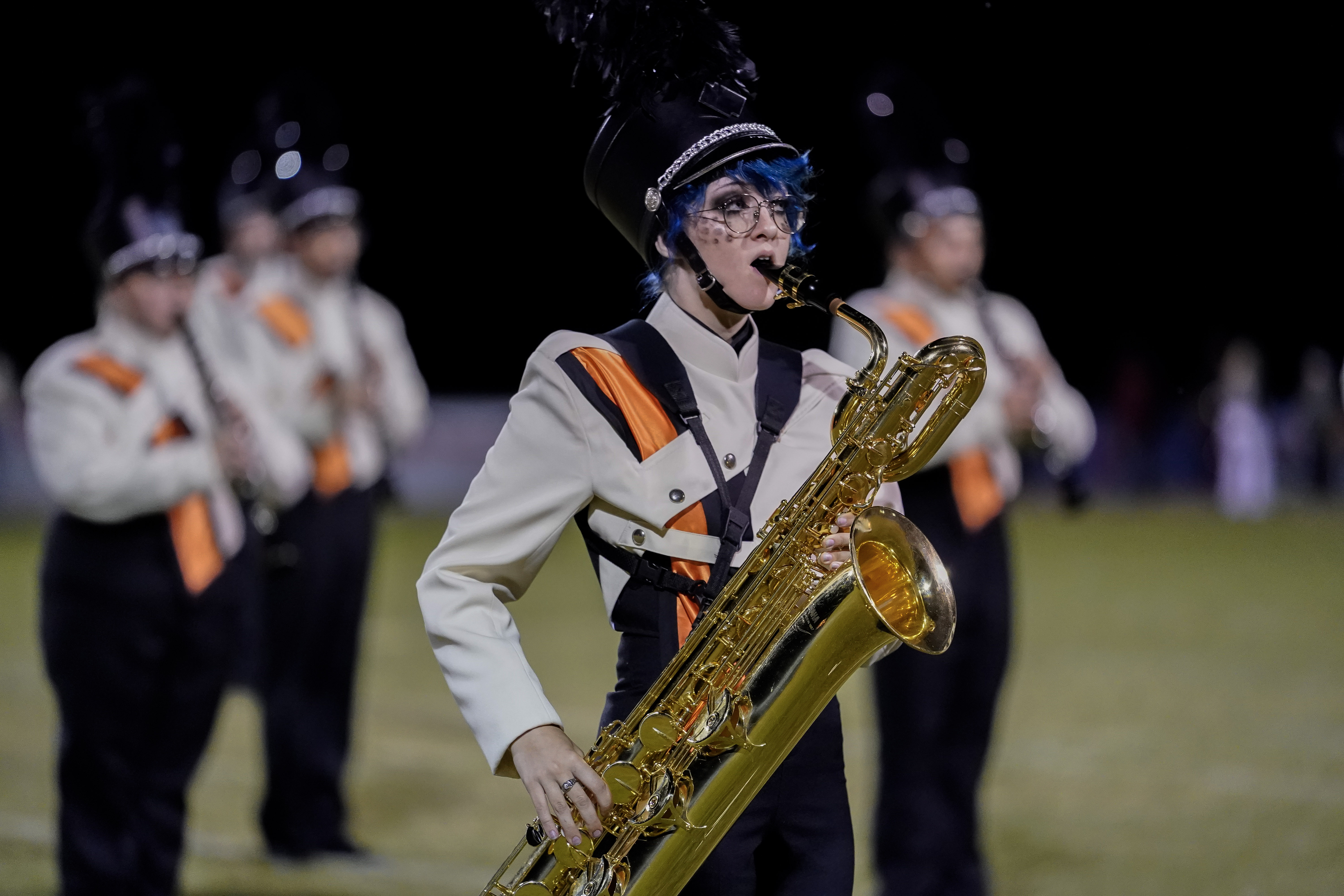 A marching band member in uniform plays a baritone saxophone on a field at night, with other band members blurred in the background.