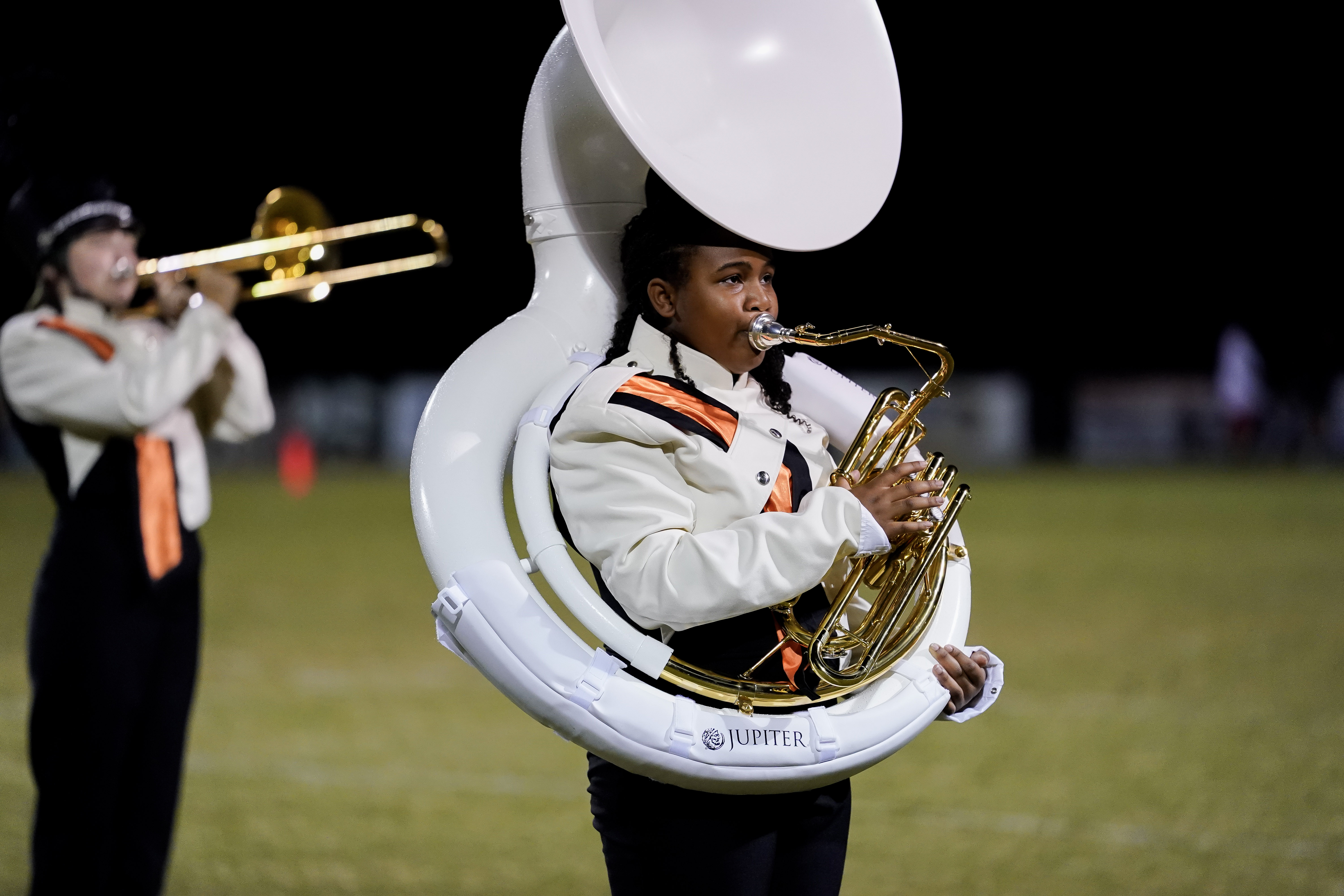 A marching band member in uniform plays a white sousaphone on a field at night, with another band member playing a trombone blurred in the background.