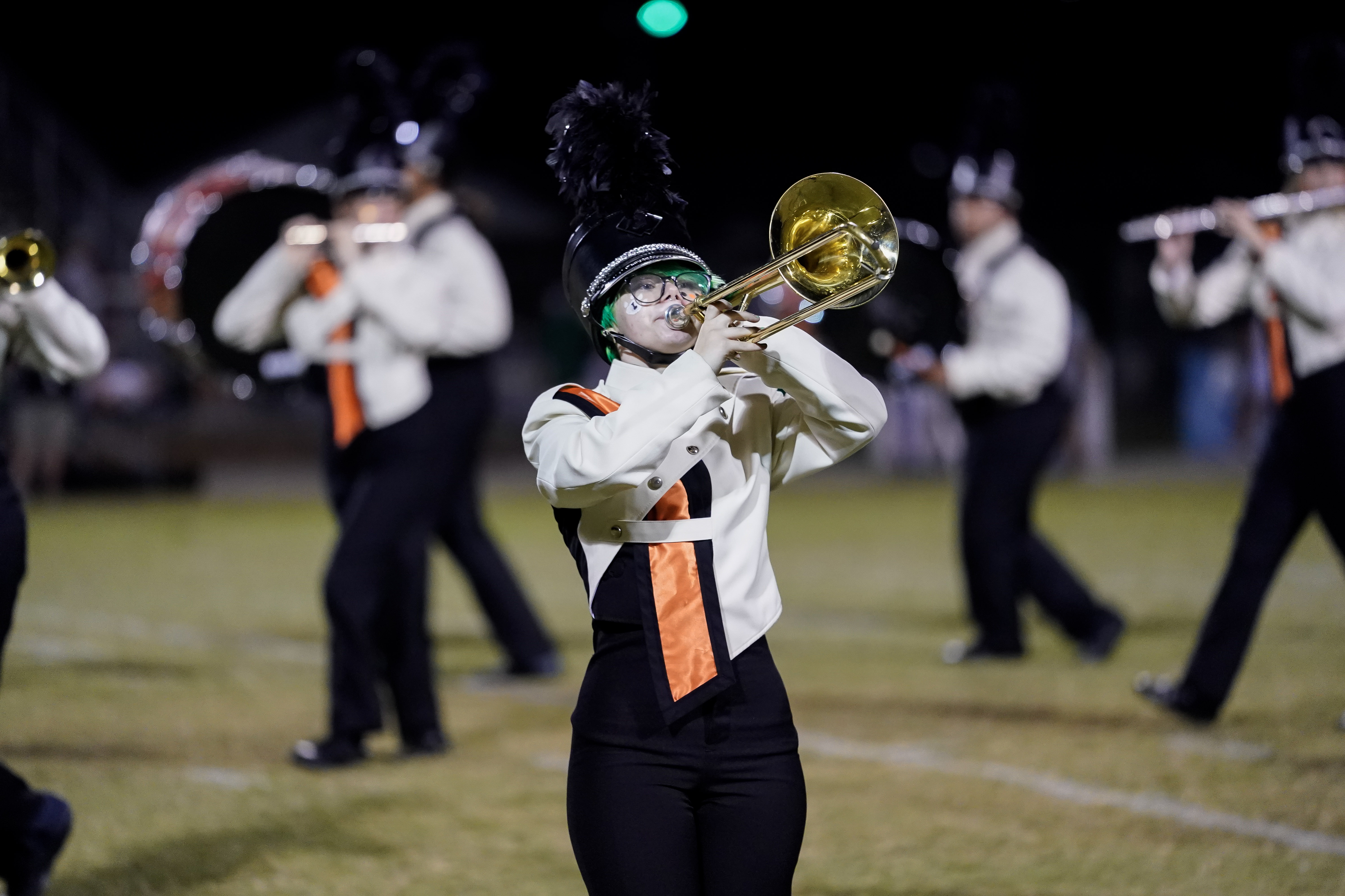 A marching band member in uniform and a feathered hat plays a trombone on a field at night, with other band members performing in the background.
