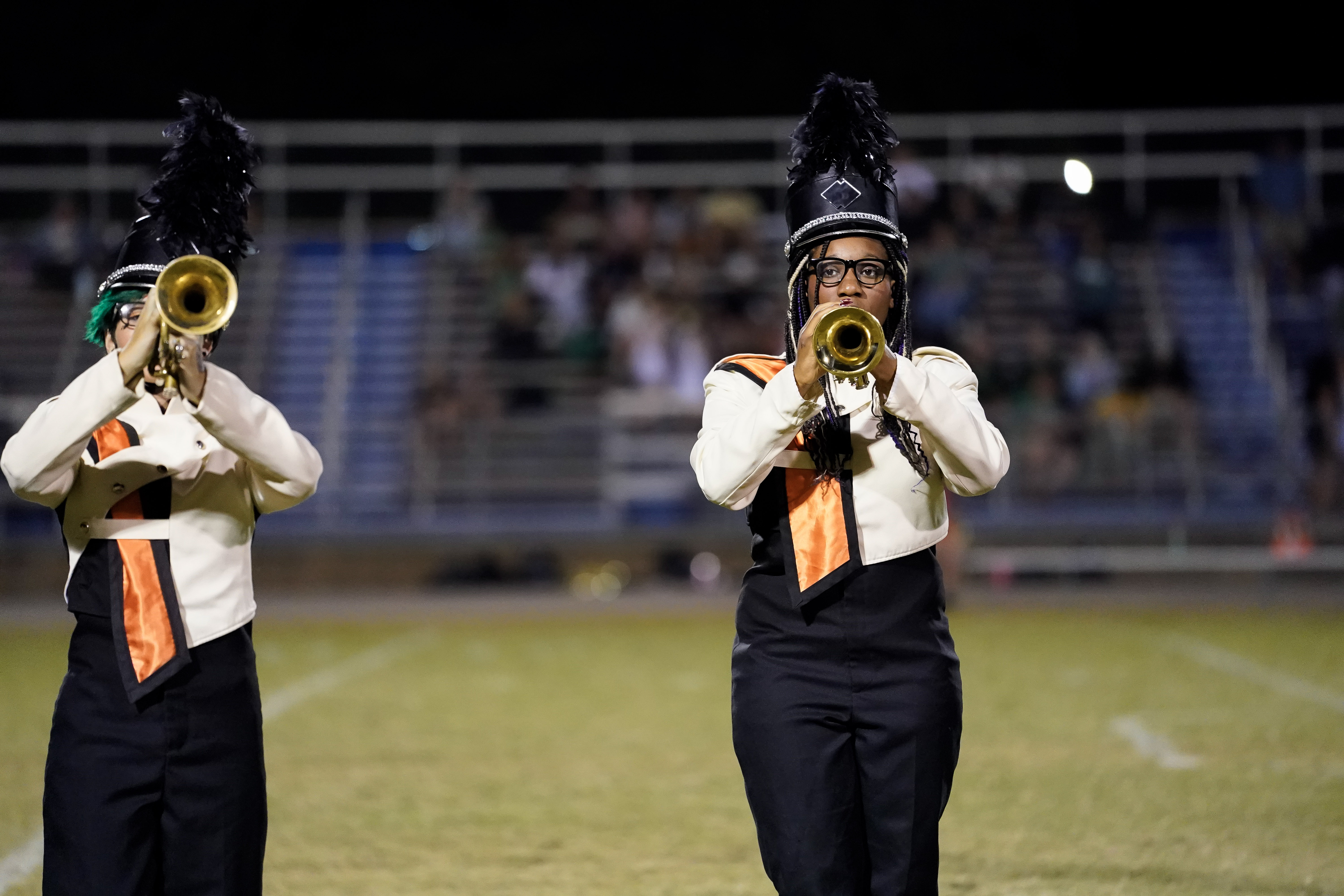 Two marching band members in black and white uniforms with feathered hats play trumpets on a football field at night; blurred bleachers with people are in the background.