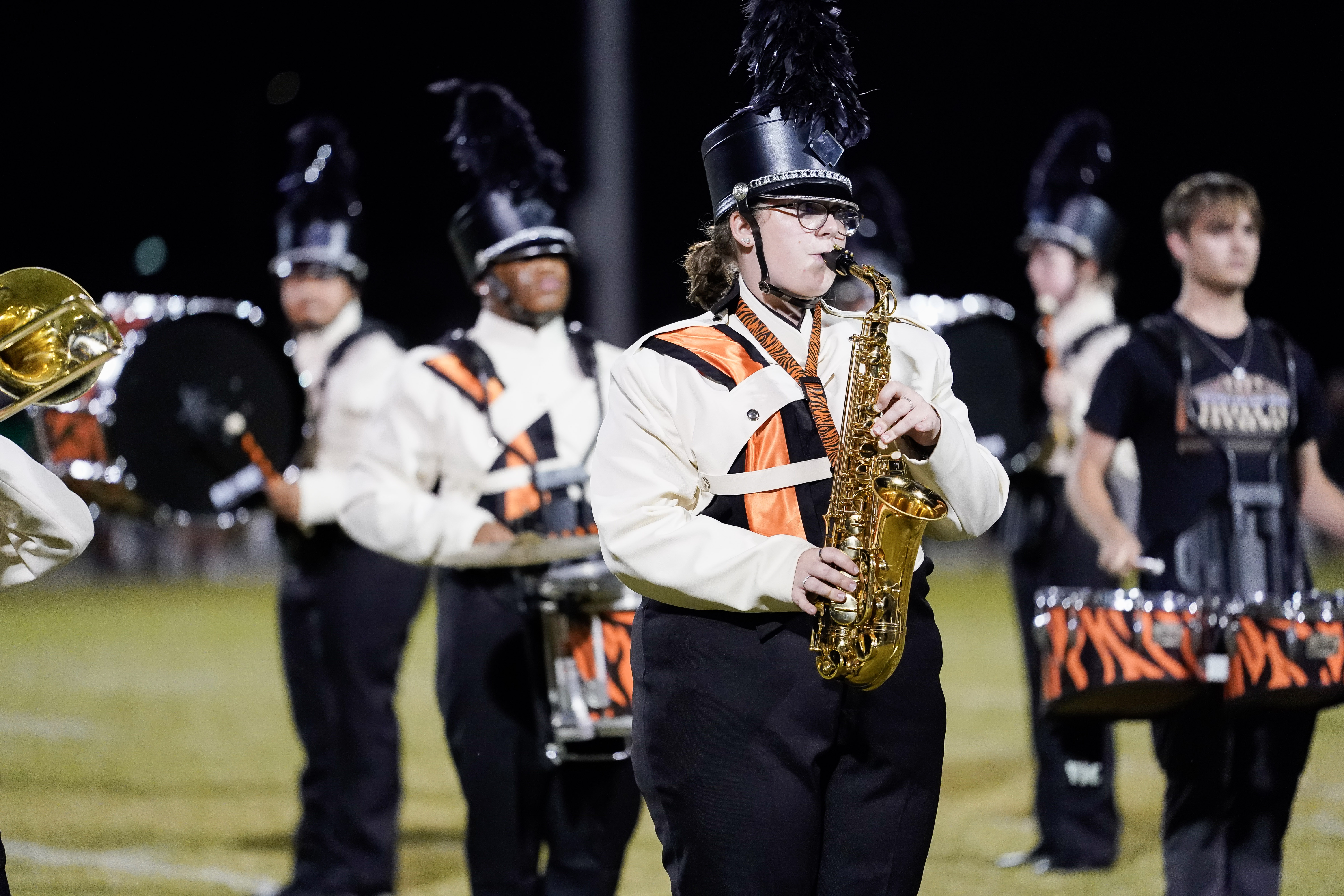 A marching band performs at night on a field. The focus is on a saxophonist in a black and white uniform with an orange stripe, wearing a tall hat with a plume. Other band members with drums are visible in the background.