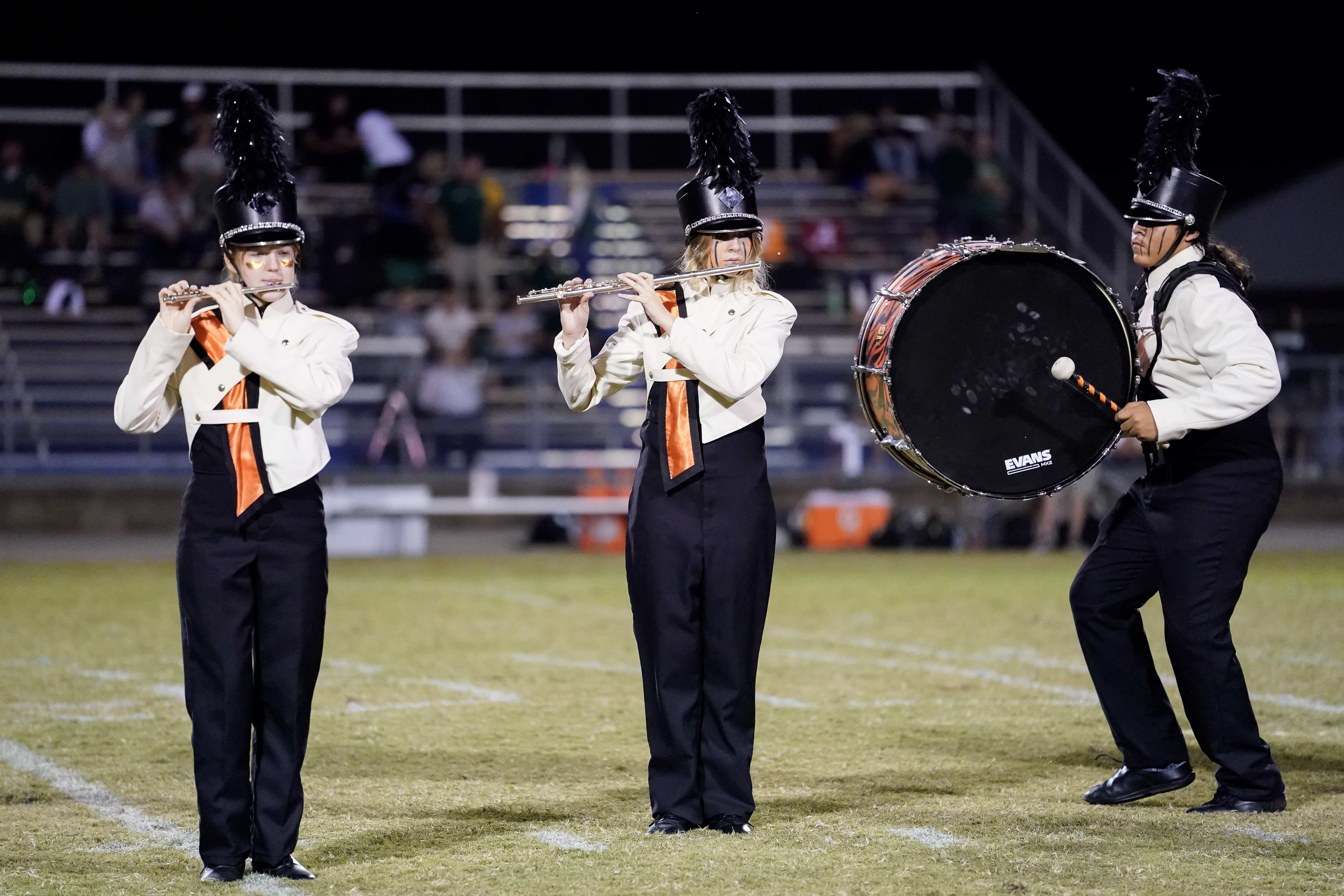 Three marching band members perform on a field at night; two play flutes while another plays a large bass drum. They wear matching uniforms with black pants, white jackets, tall hats, and orange ties. Bleachers with people are in the background.