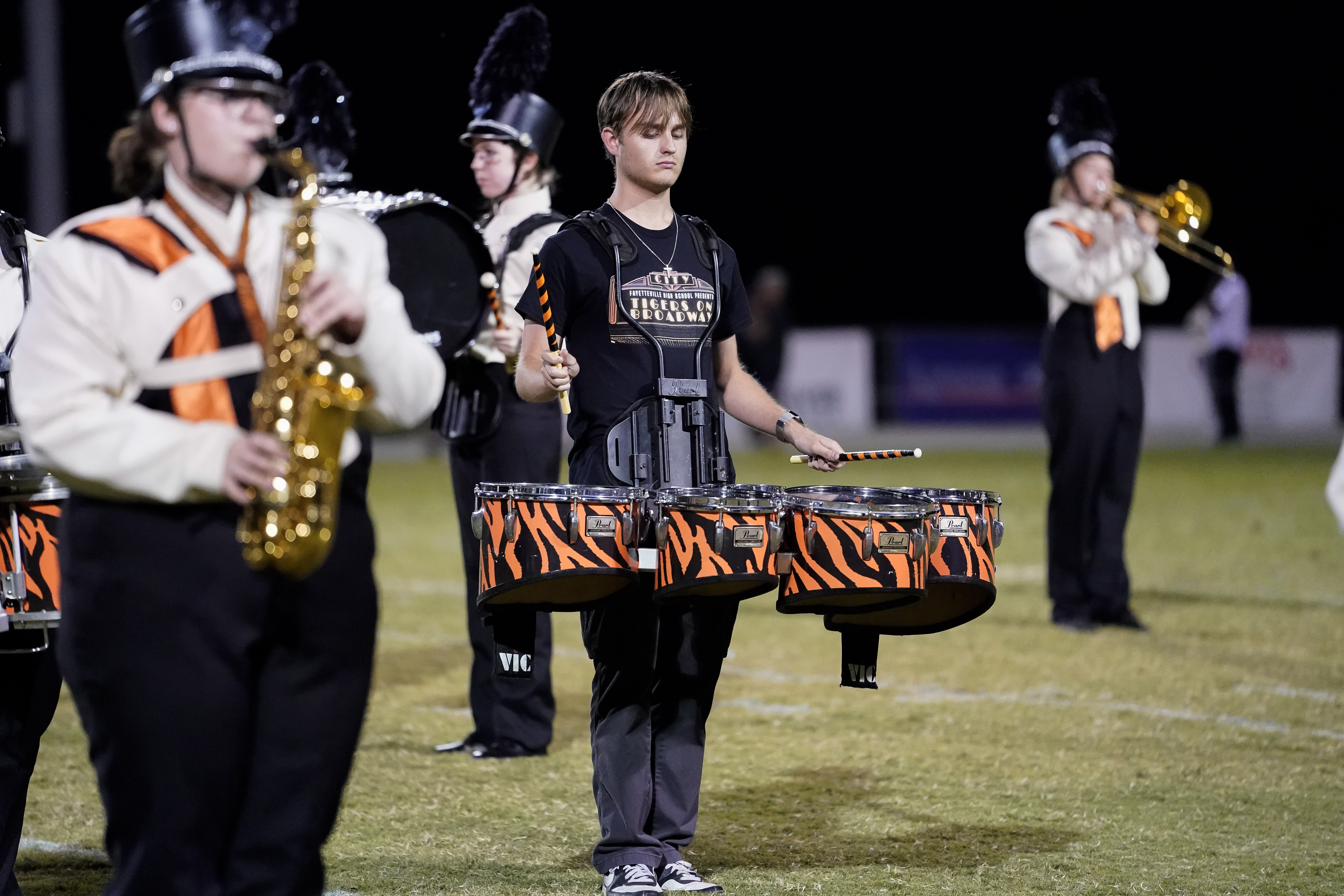 A high school marching band drumline performs on a football field at night, with members wearing black and orange uniforms and hats featuring black plumes. One member in a black t-shirt carries tenor drums.
