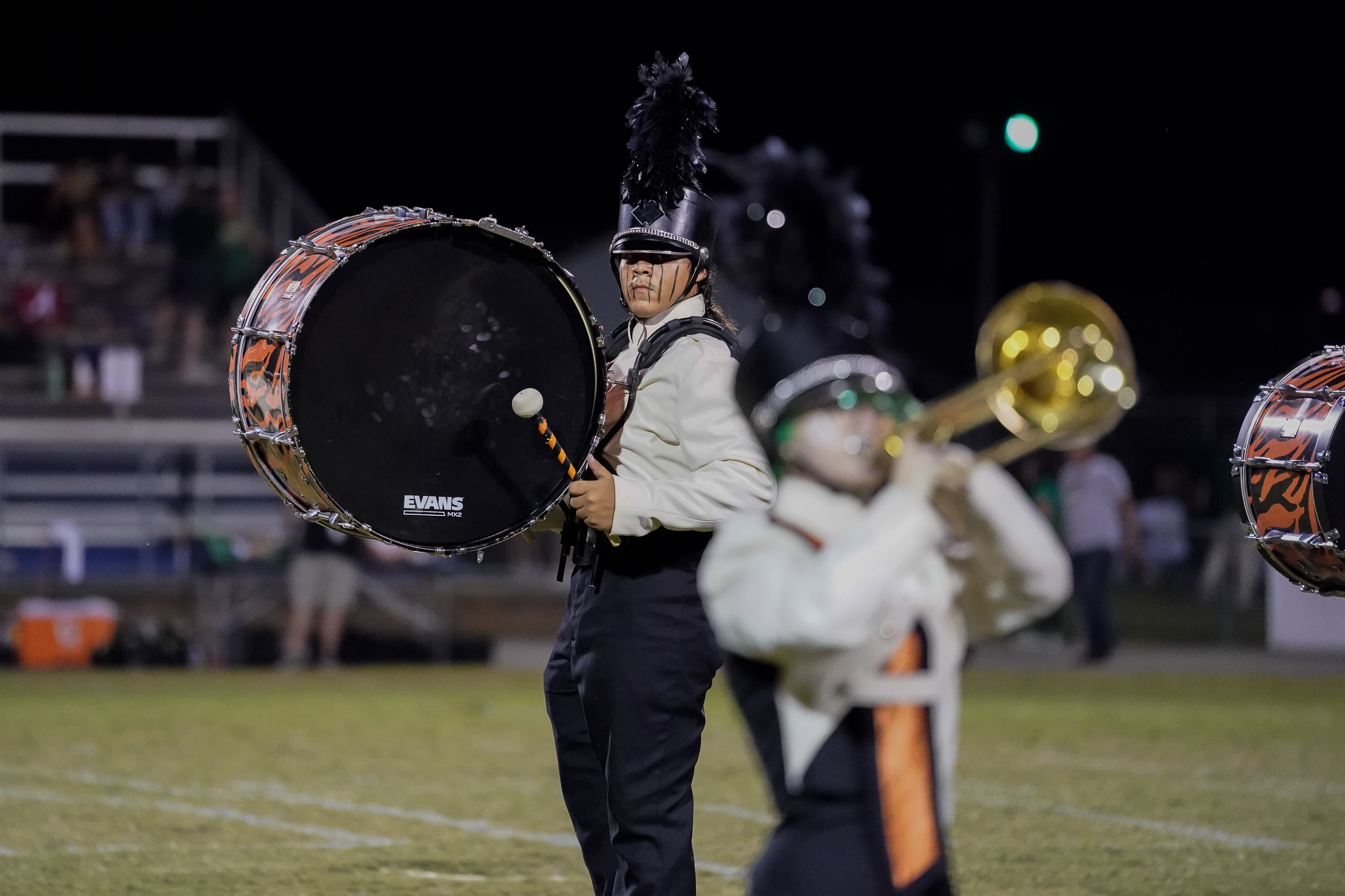 A marching band member in uniform plays a large bass drum on a field at night, while another member in the foreground plays a brass instrument. The background shows blurred bleachers and spectators.