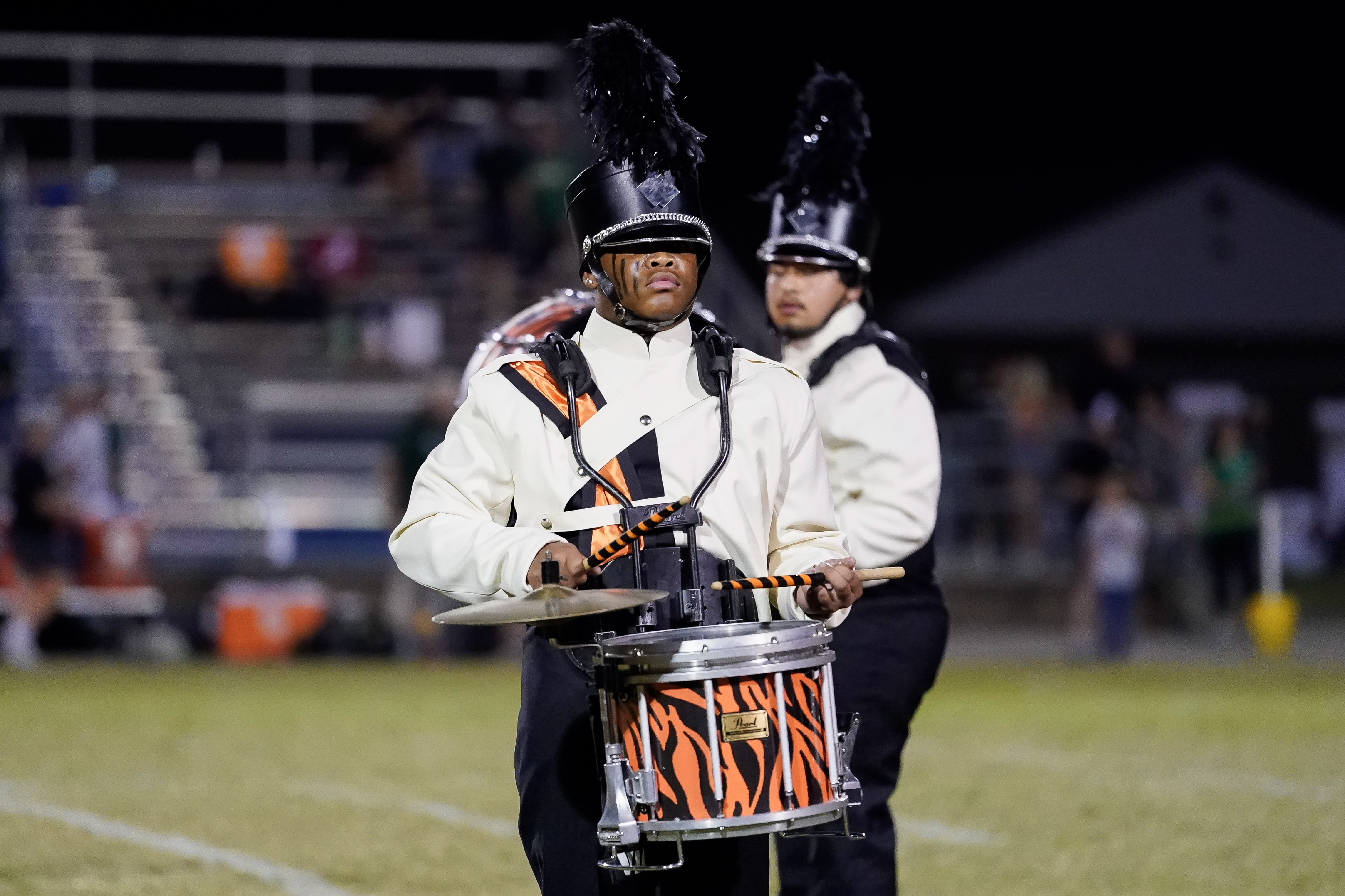 A marching band member in uniform plays a snare drum with an orange and black tiger-stripe design on a football field at night; another band member is visible in the background.